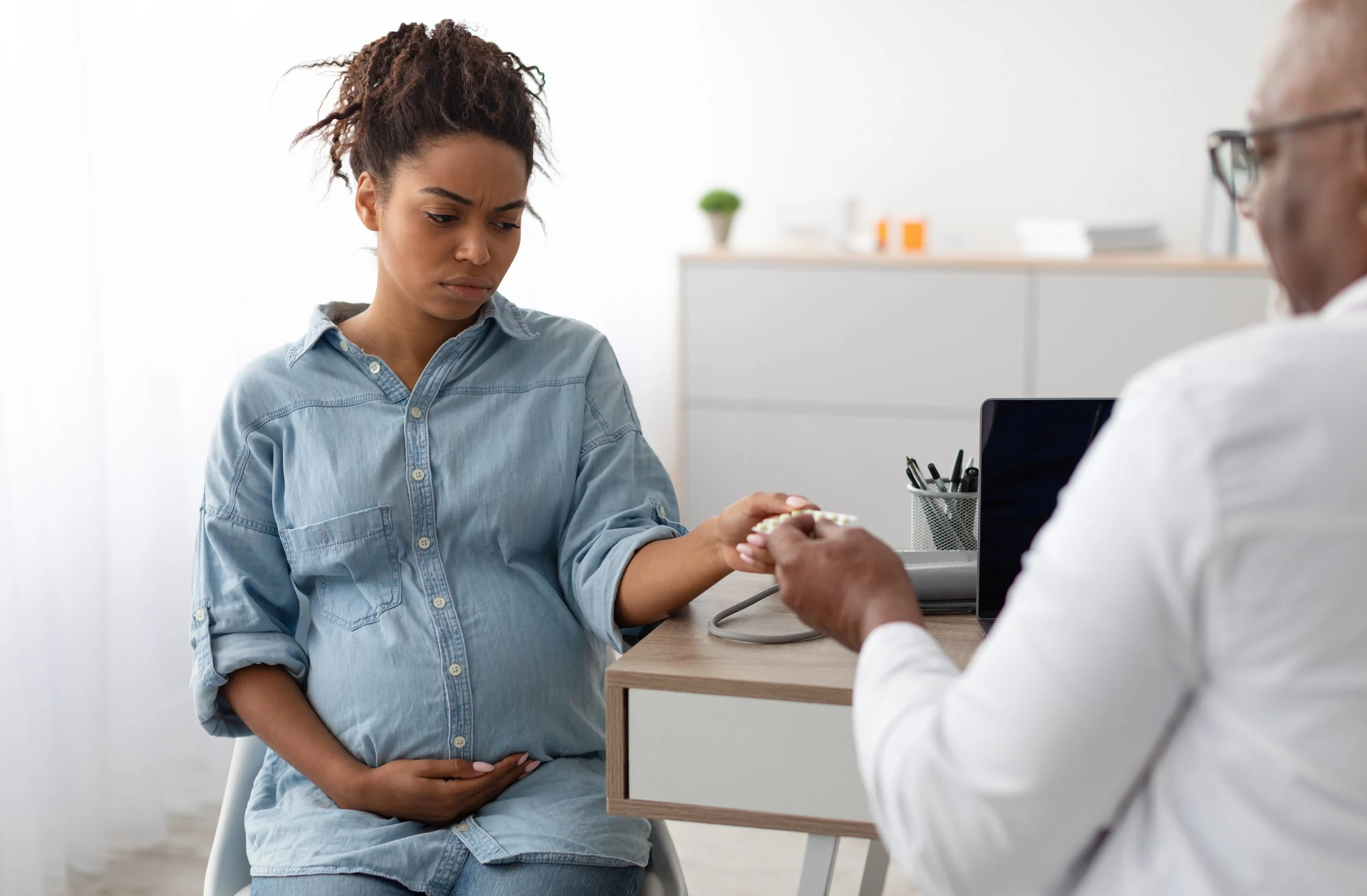 Young woman sitting in a doctor's office, holding her stomach, looking concerned while a doctor hands her medication.