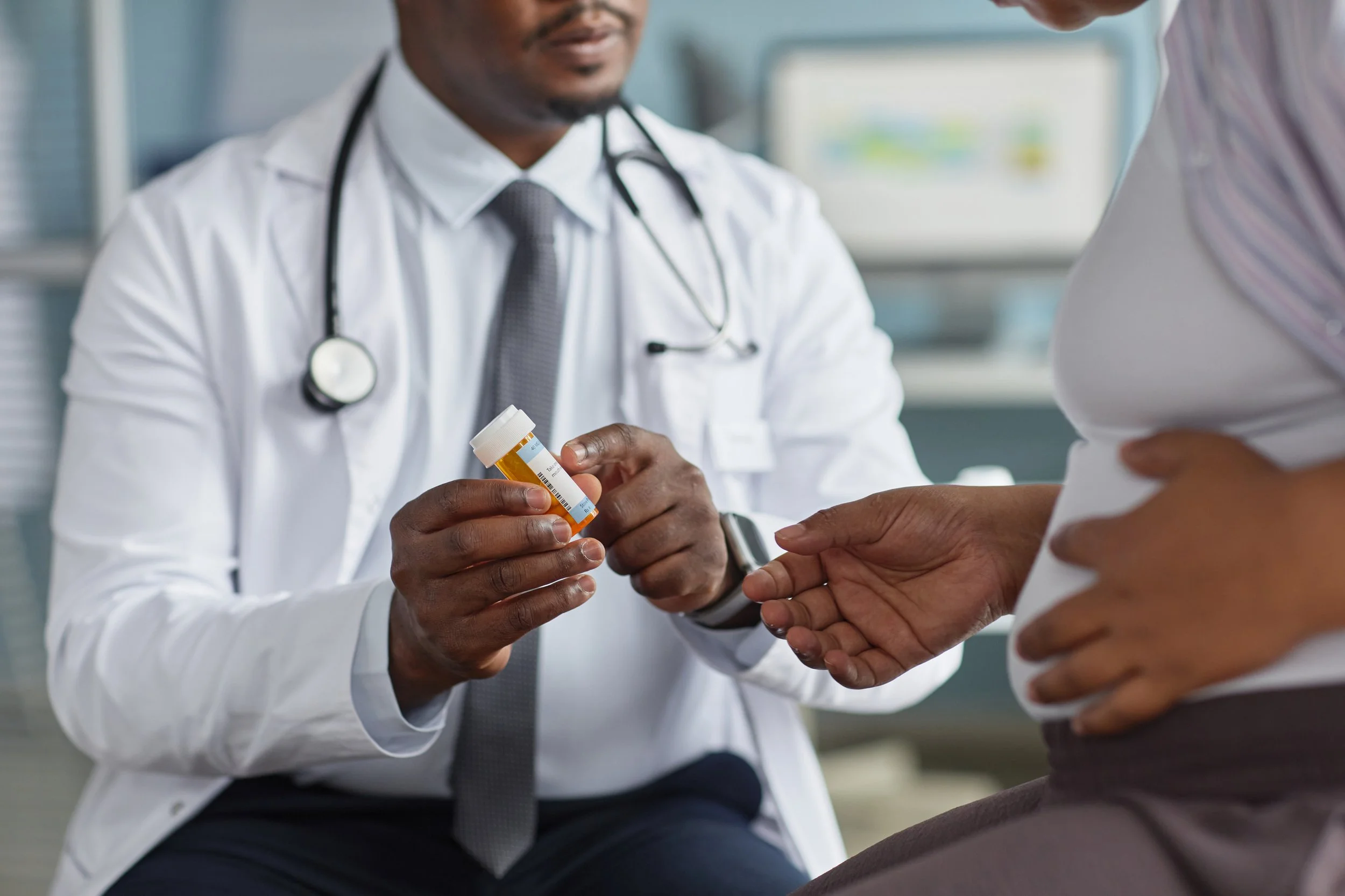 A doctor in a white coat with a stethoscope around his neck handing a prescription medication bottle to a patient in a clinical setting.