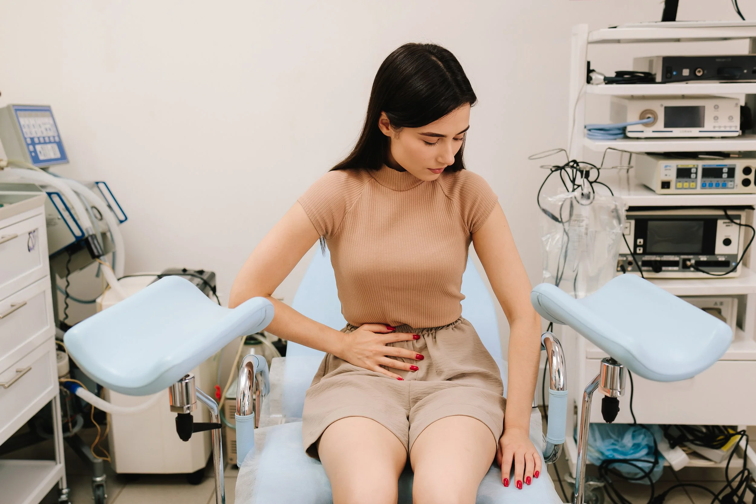 A young woman with black hair, wearing a beige top and shorts, sitting on a medical examination table with her hand on her stomach in a clinical setting. Medical equipment and monitors are visible in the background.