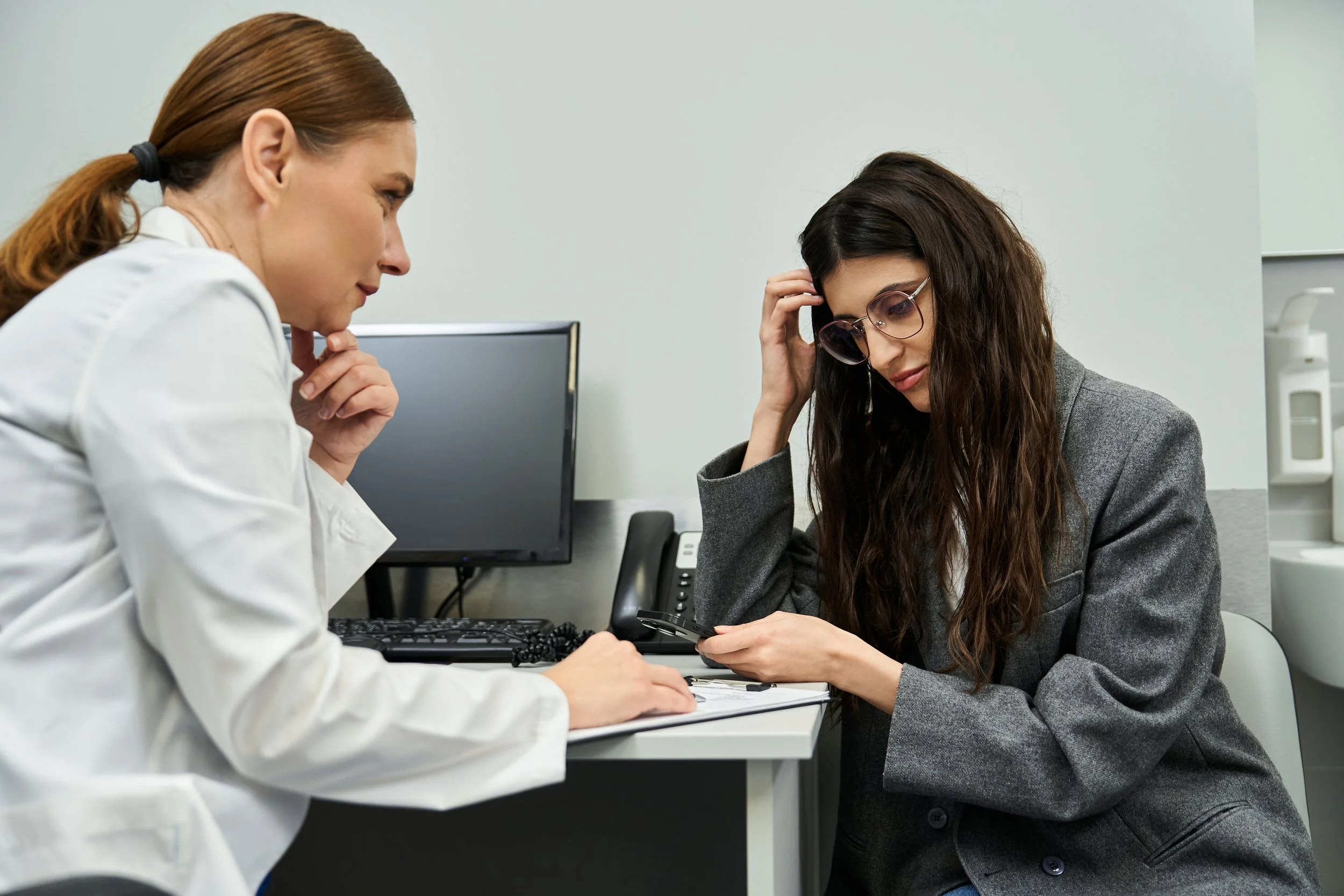 A woman with long curly hair and glasses sitting at a desk, looking down at her phone, while a female medical professional in a white coat stands beside her, holding a clipboard and discussing something. The scene takes place in a medical office or clinic with a computer monitor and phone on the desk.