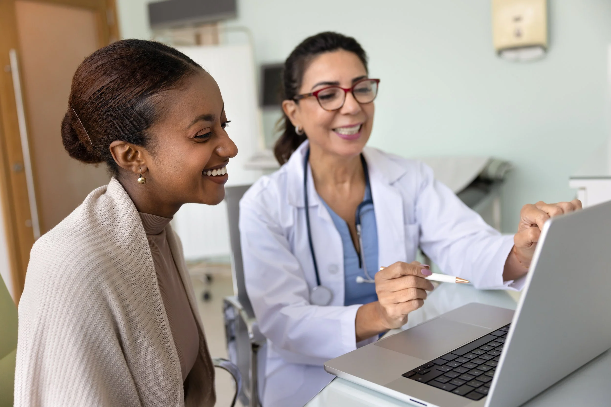A healthcare professional and a woman looking at a laptop in a clinical setting, both smiling.