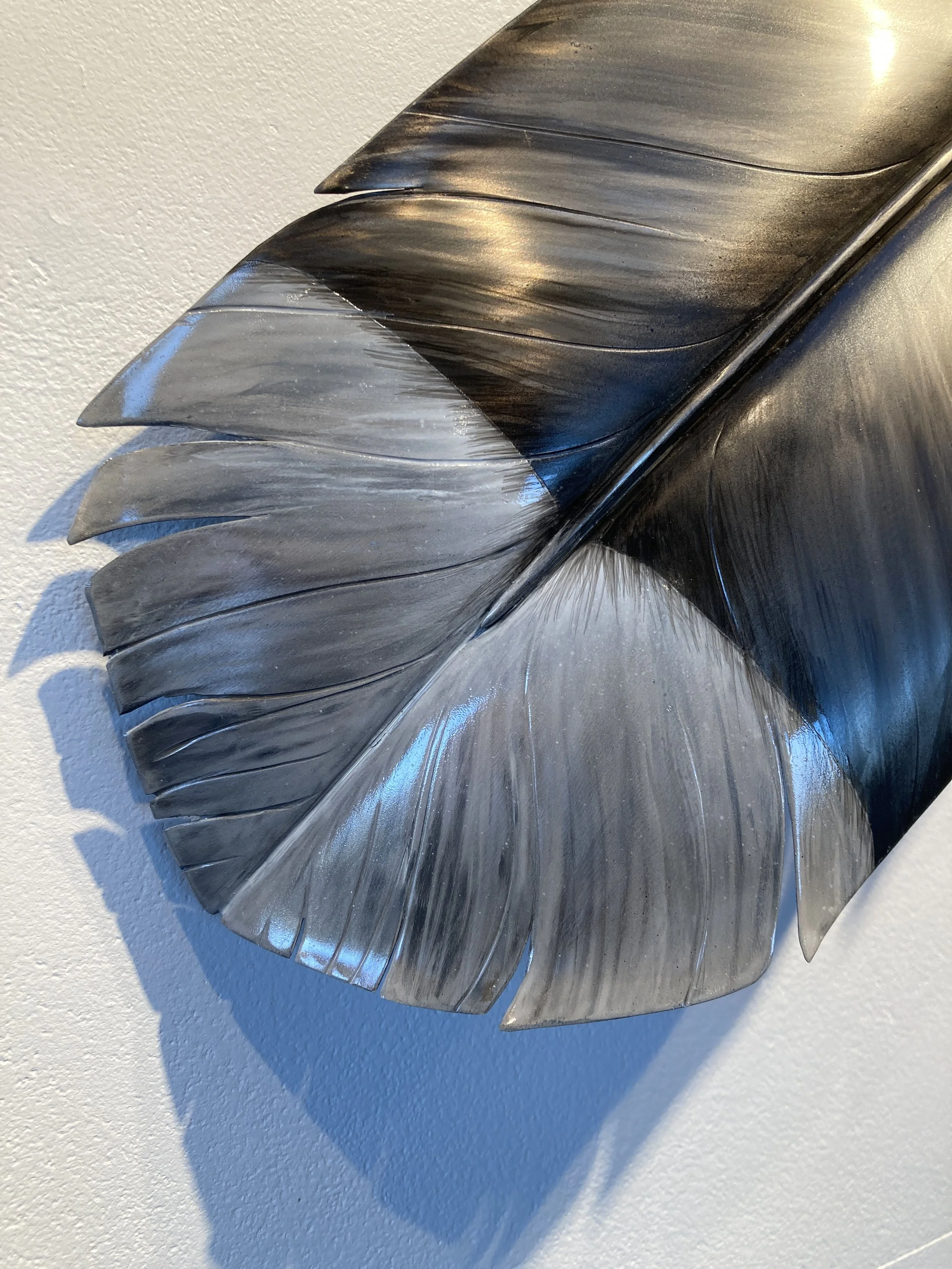 Close-up of a metallic sculpture resembling a feather with detailed ridges, mounted on a white wall, casting a shadow.