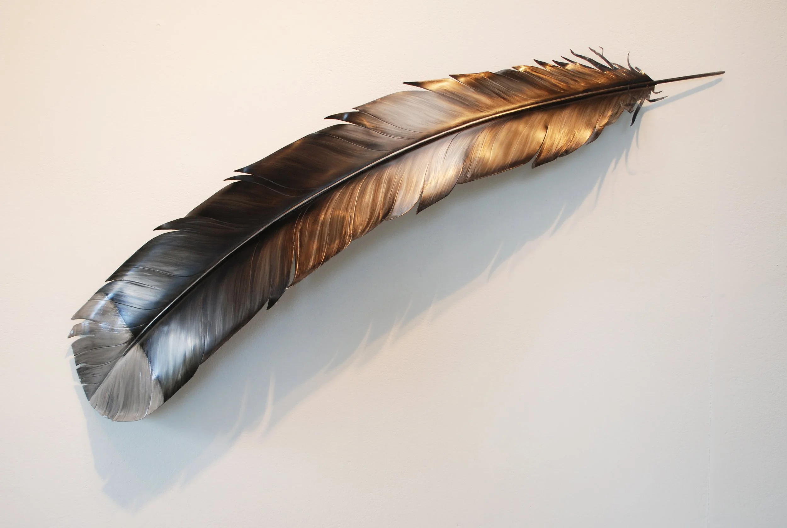 Decorative wall art of a large, metallic feather mounted on a plain wall.