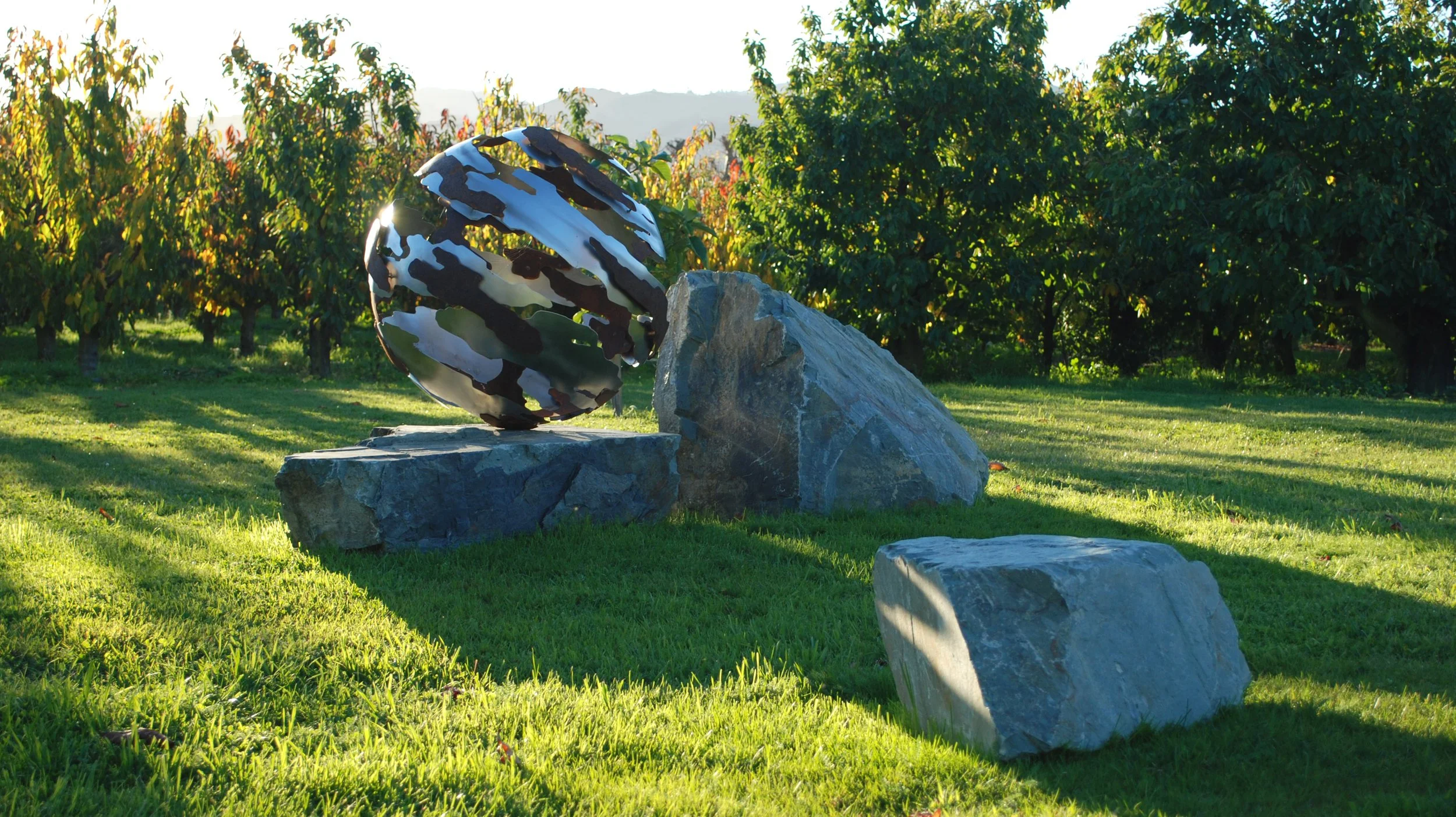 Outdoor sculpture featuring a mosaic metal sphere balanced on large rocks in a grassy area with orchard trees in the background