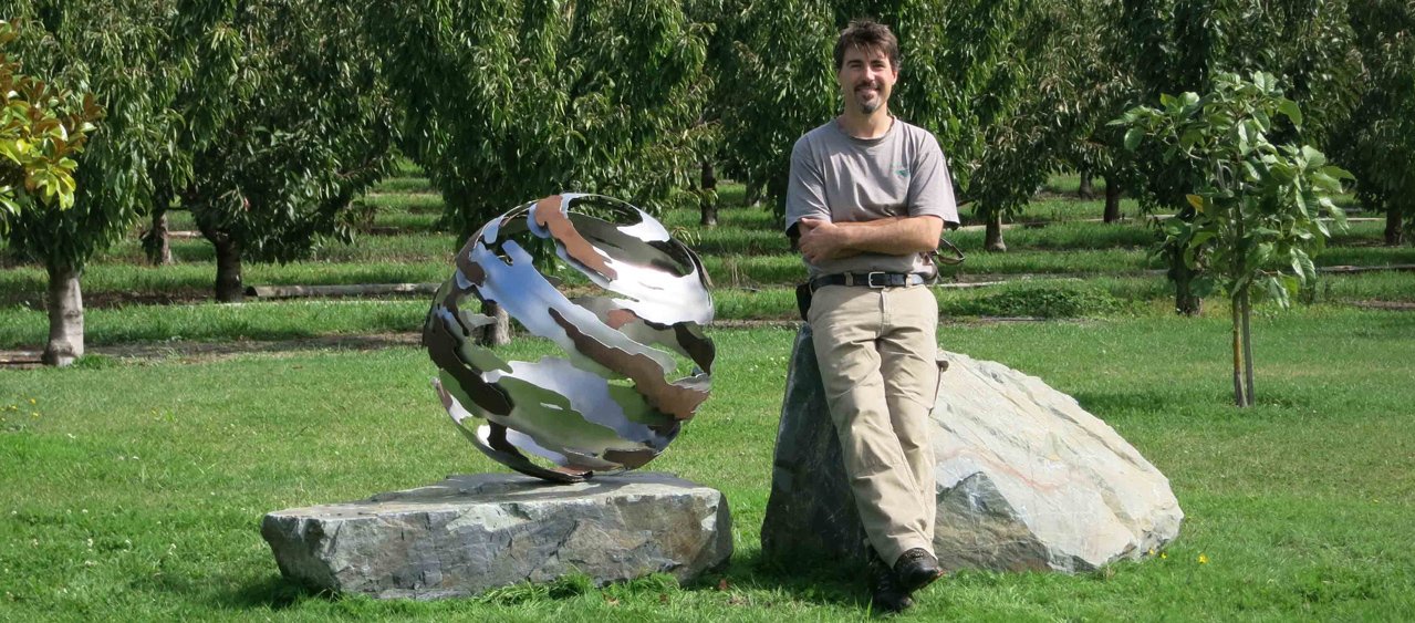 A man with a beard and gray T-shirt stands with arms crossed, leaning against boulder and next to a spherical metal sculpture, in a grass field of an orchard.