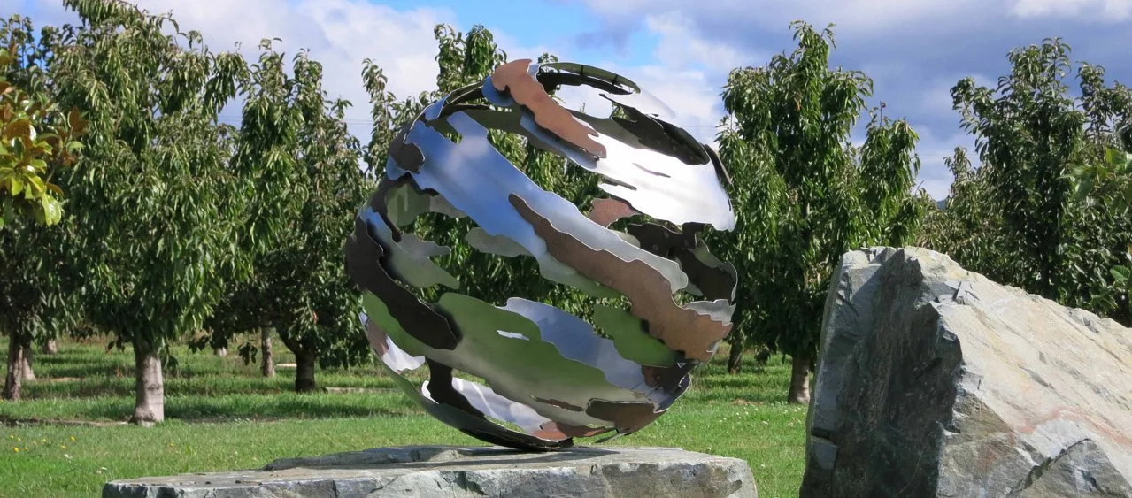 Metal sculpture of a globe with swirling bands, situated outdoors among trees and large rocks.