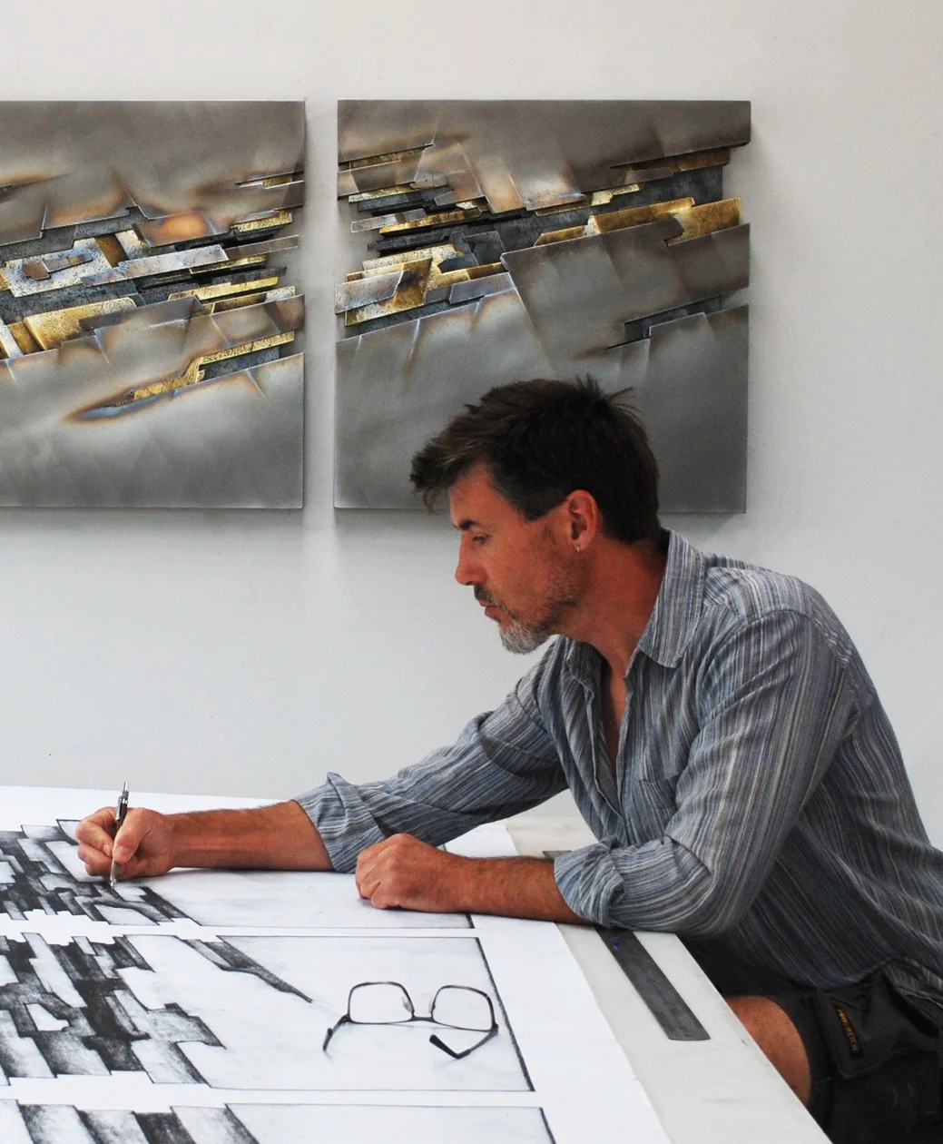 A man with dark hair, a gray beard, wearing a gray striped shirt, sitting at a table, sketching architectural plans or drawings with a black pen, with an artwork on the wall behind him.