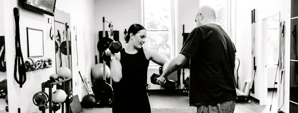 A woman lifting dumbbells while a trainer assists in a gym.