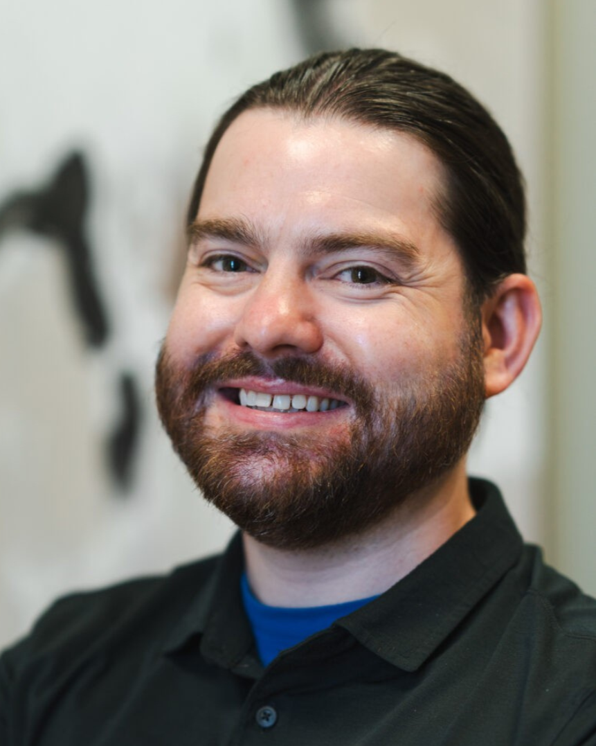 A smiling man with dark hair and a beard, wearing a black shirt, standing indoors with abstract wall art in the background.