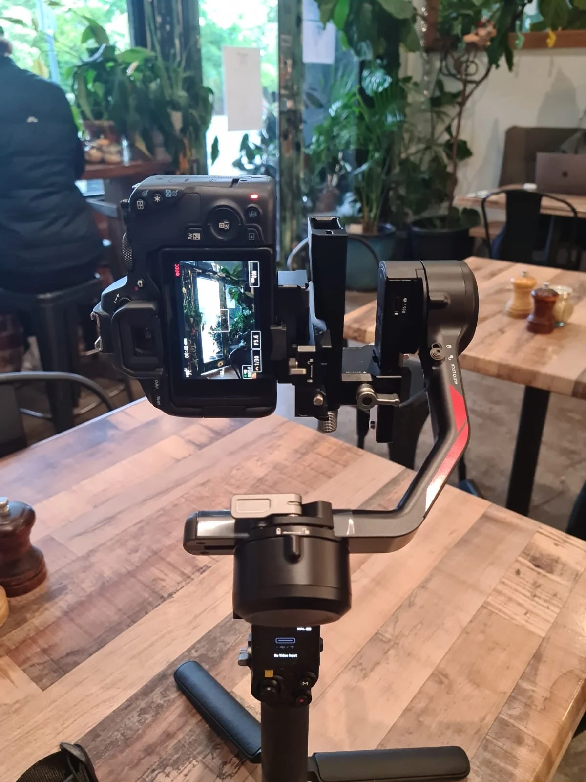 A camera mounted on a gimbal stabilizer recording a shot of a table in a café or restaurant with wooden tables, salt and pepper shakers, and plants in the background.