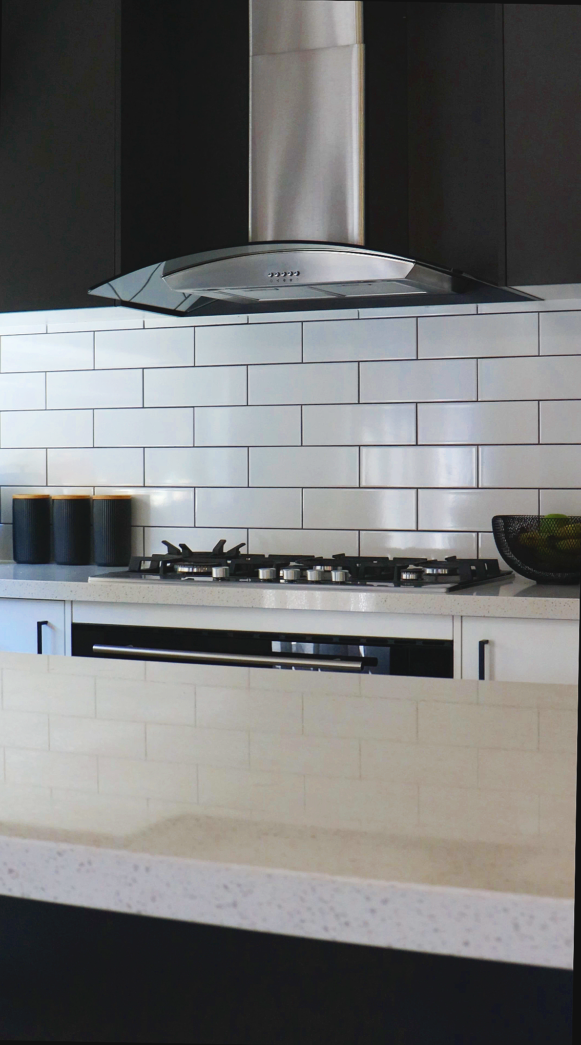 Modern kitchen with black and white cabinetry, stainless steel range hood, gas stove, and white subway tile backsplash. Black canisters and a black bowl with green produce are on the countertop.