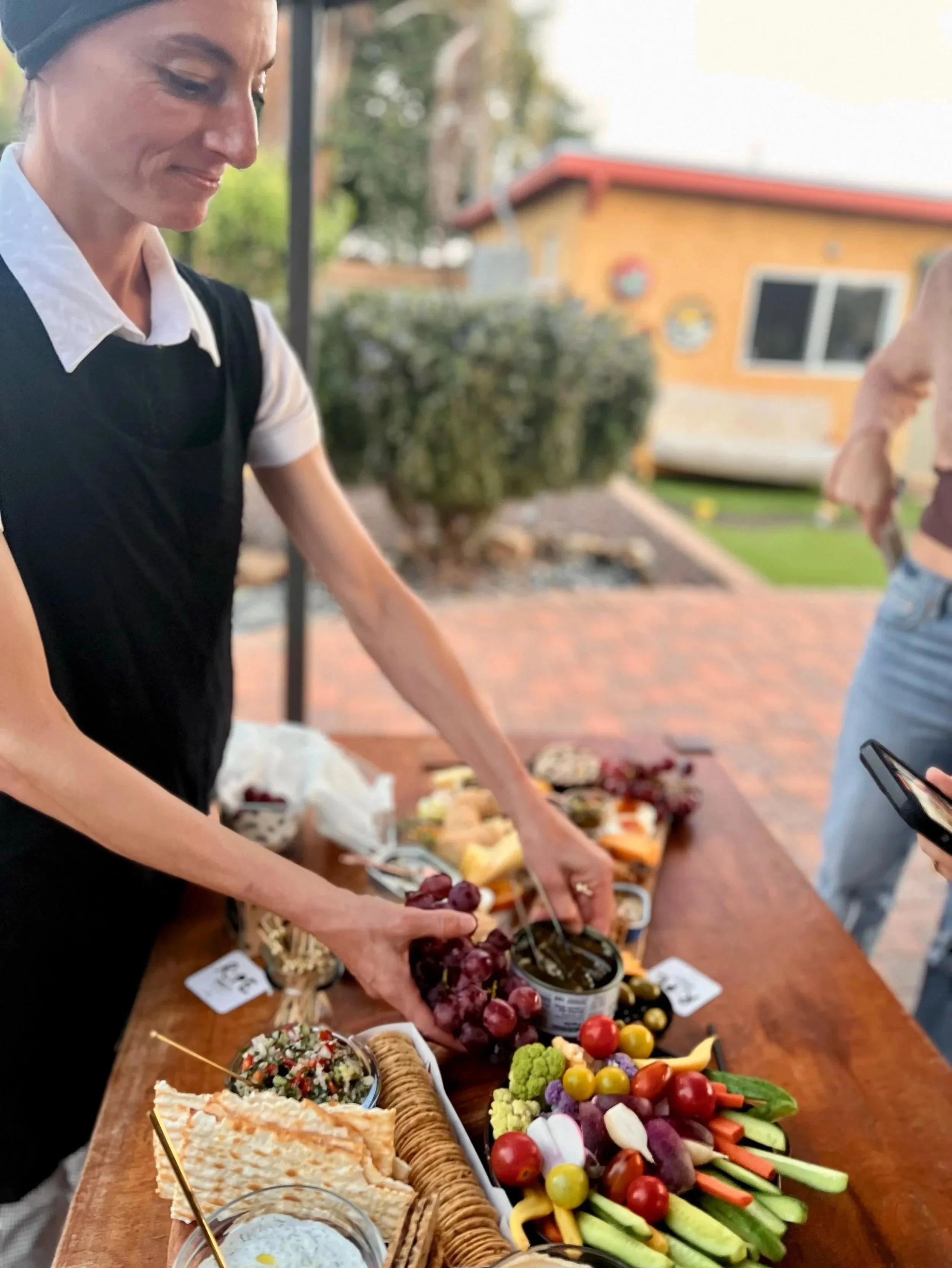 A woman in a black apron and white shirt reaching for grapes on a table filled with cheese, crackers, and fresh vegetables outdoors.