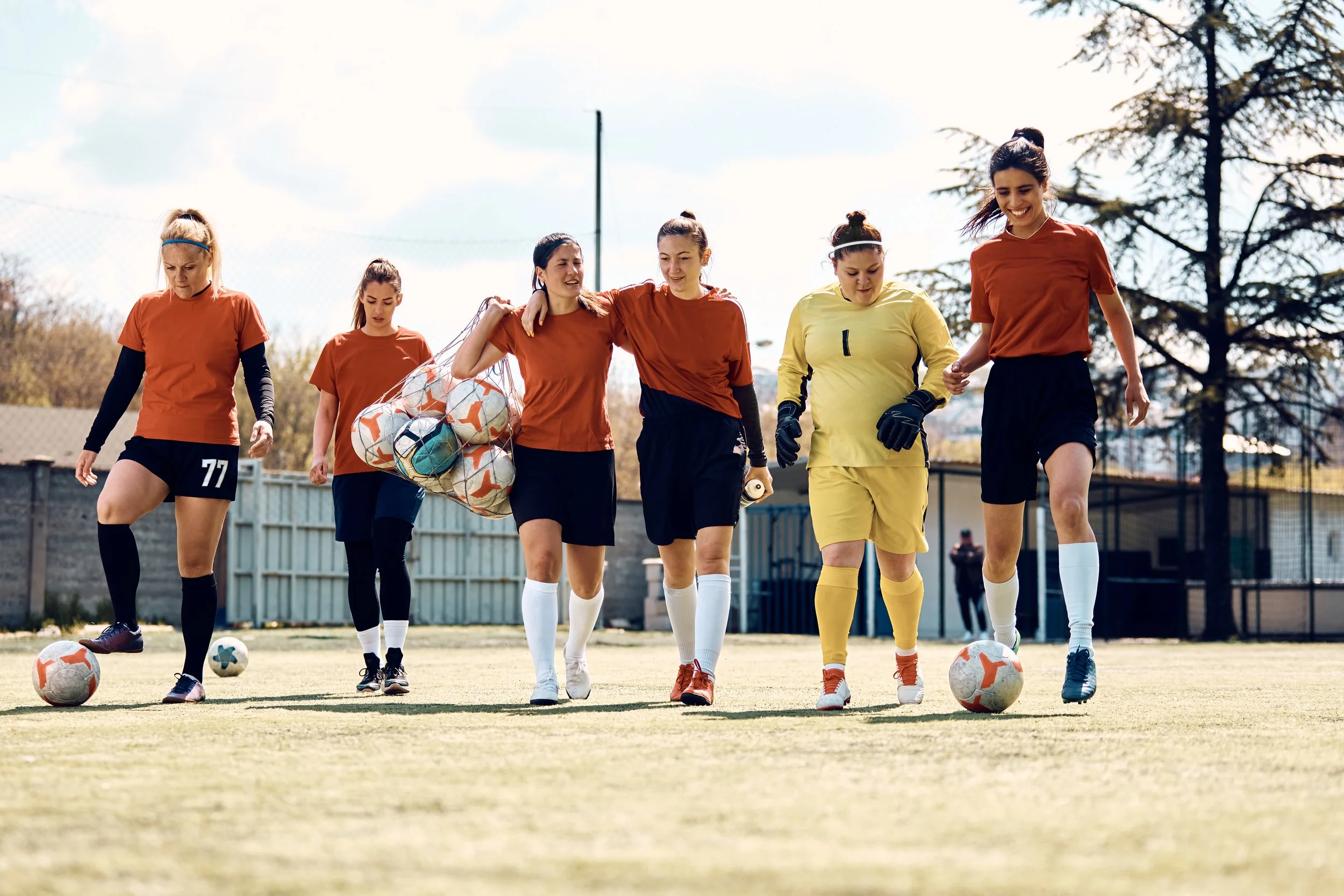 Six women, some in soccer uniforms, walking on a soccer field with soccer balls and a net in the background on a sunny day.