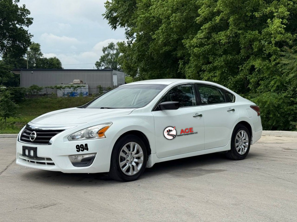 White sedan with a logo and number on the side, parked on a concrete lot with green trees in the background.