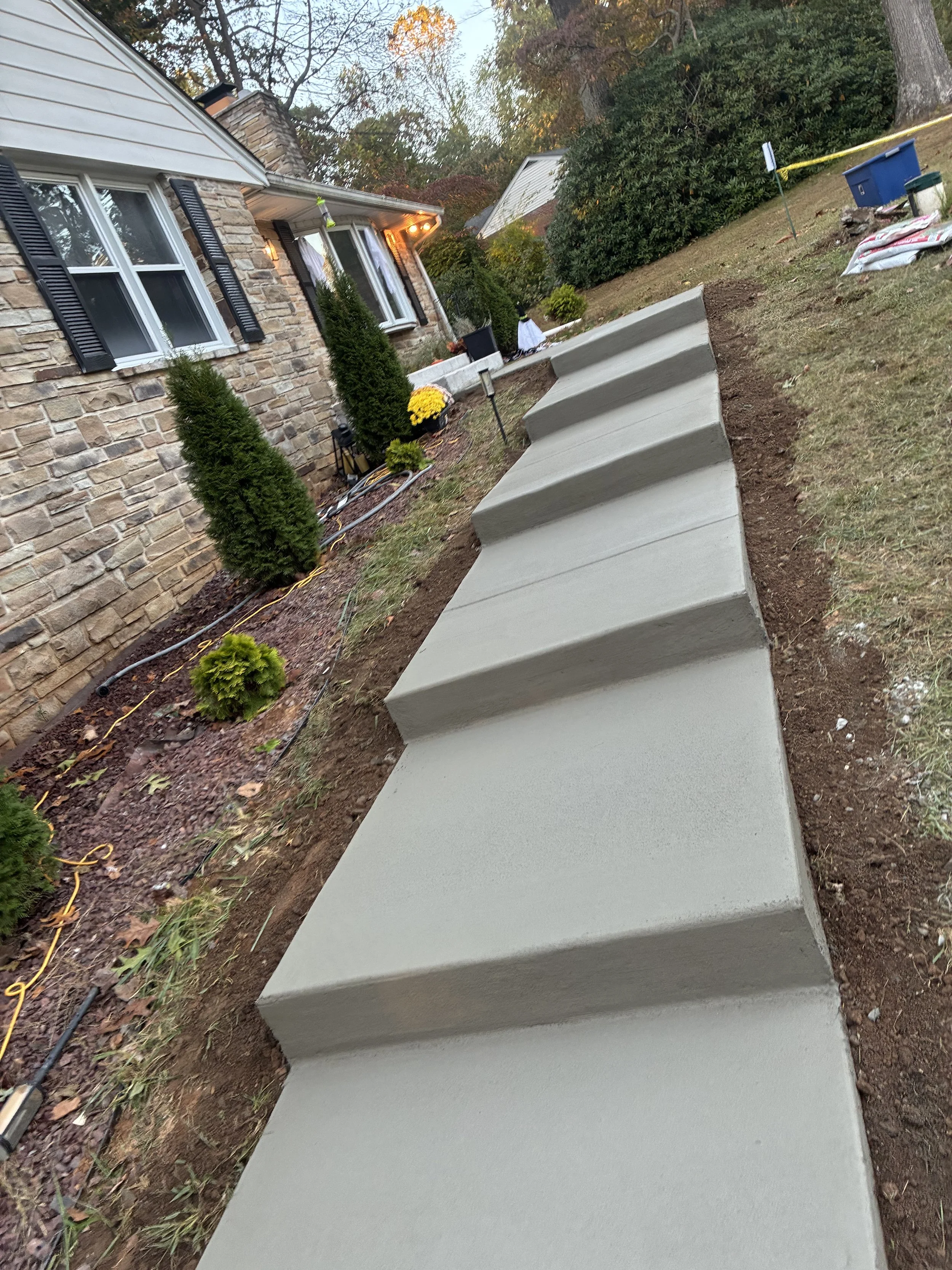 Newly poured concrete sidewalk in a residential yard, with a brick house on the left and plants along the walkway, evening lighting, and yardwork tools.