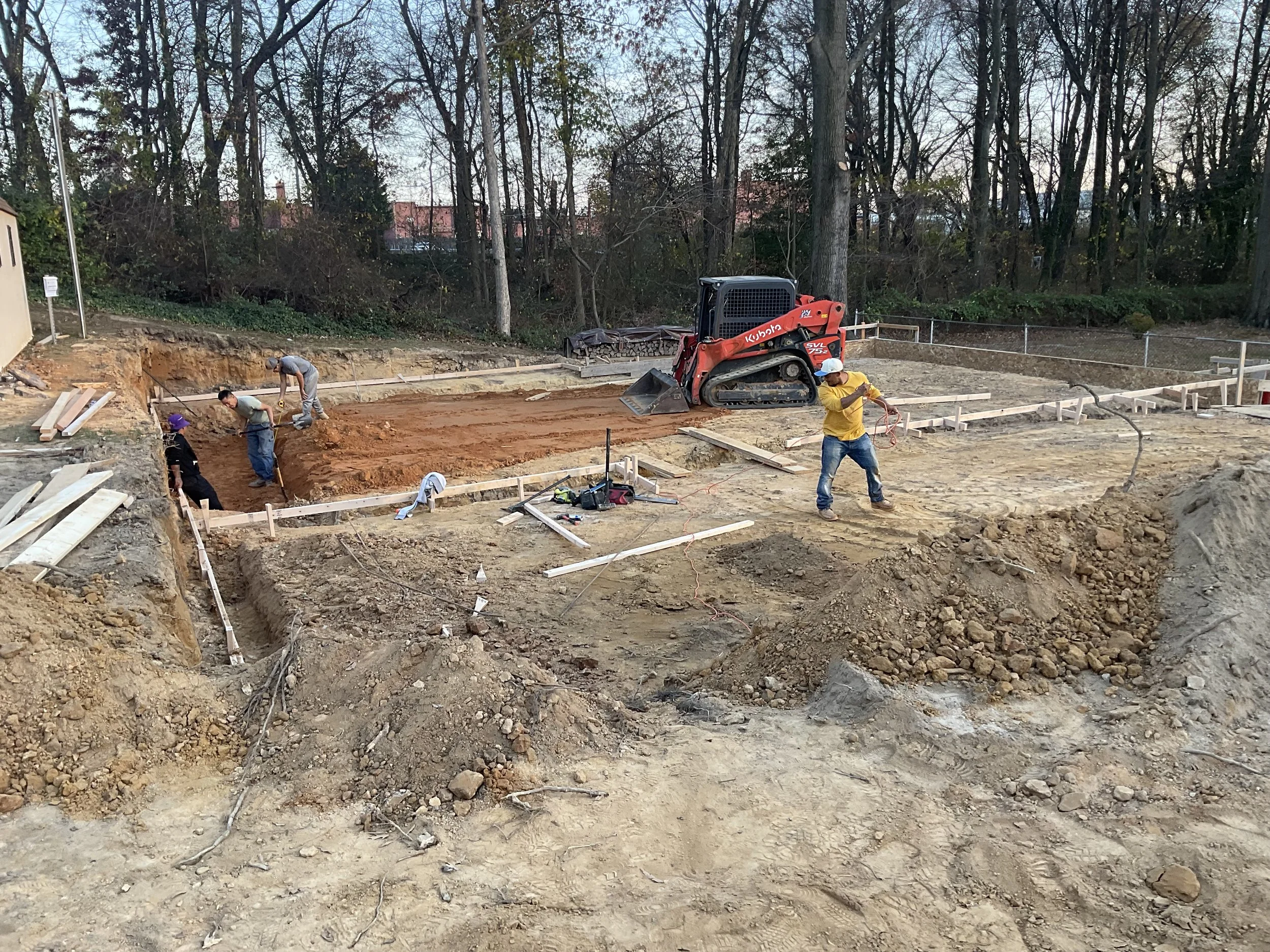 Construction site with workers preparing the ground, surrounding trees, and a small red tracked excavator.