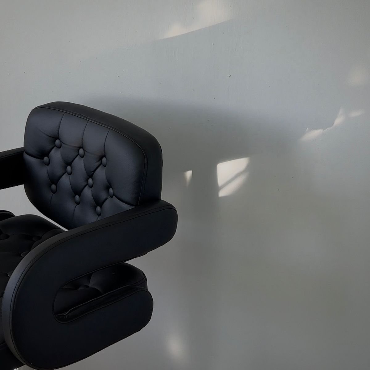 Part of a black office chair with tufted leather upholstery, casting a shadow on a white wall.