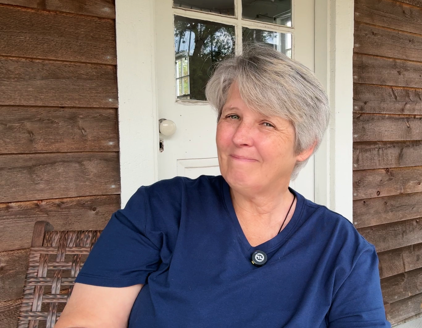 A woman with short gray hair, wearing a dark blue shirt and a round pendant necklace, smiling while sitting outdoors on a wooden bench next to a white door with glass windows. The background includes a wooden wall and greenery reflected in the window.