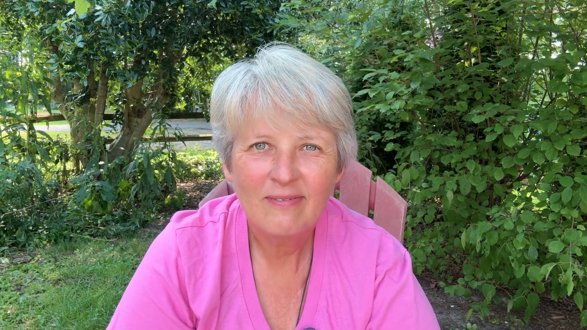 A middle-aged woman with short gray hair and blue eyes, wearing a pink shirt, sitting outdoors on a pink wooden chair surrounded by green foliage and trees.