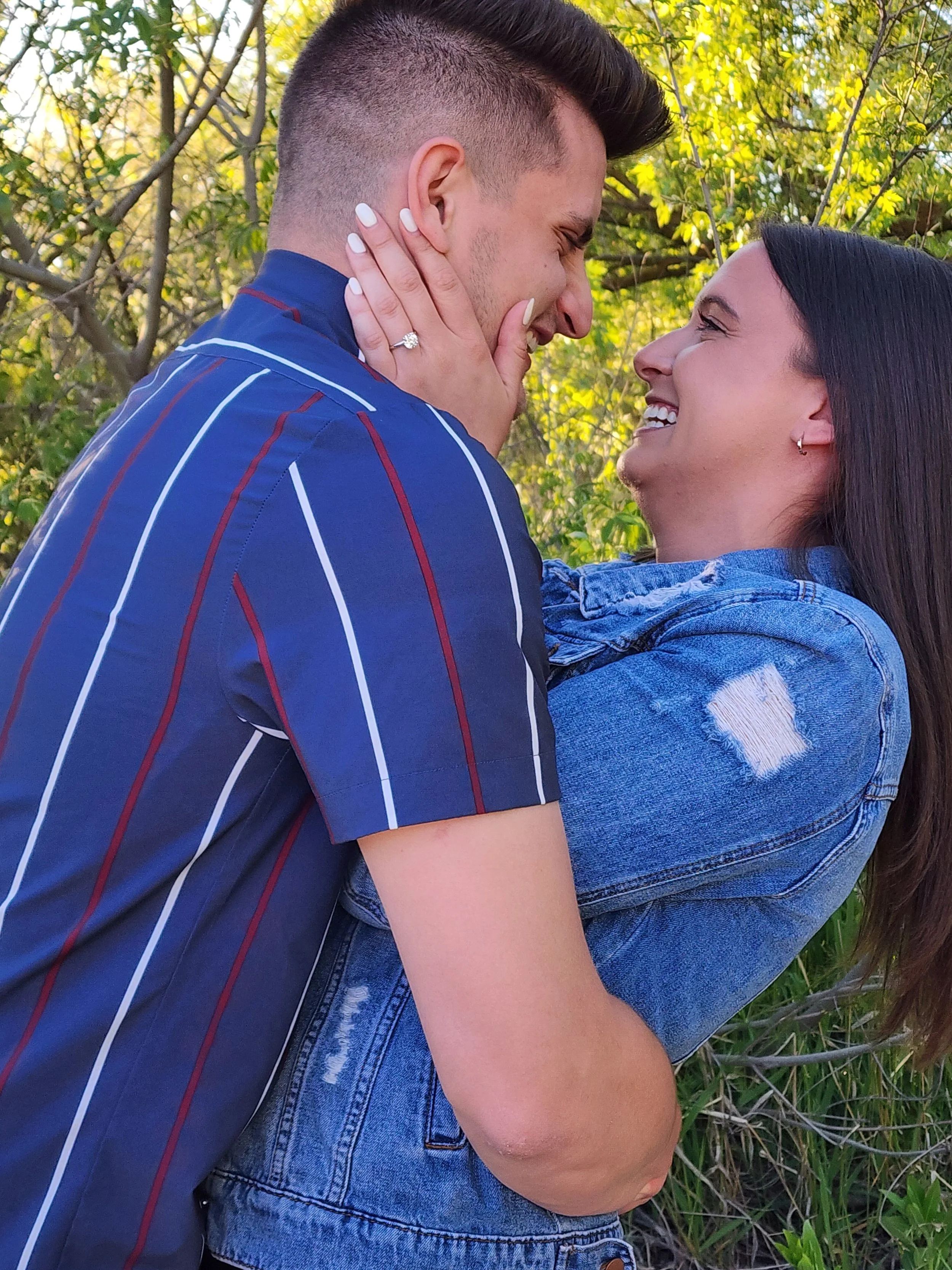 A happy couple embracing outdoors near green trees, with the woman holding the man's face gently and both smiling at each other.