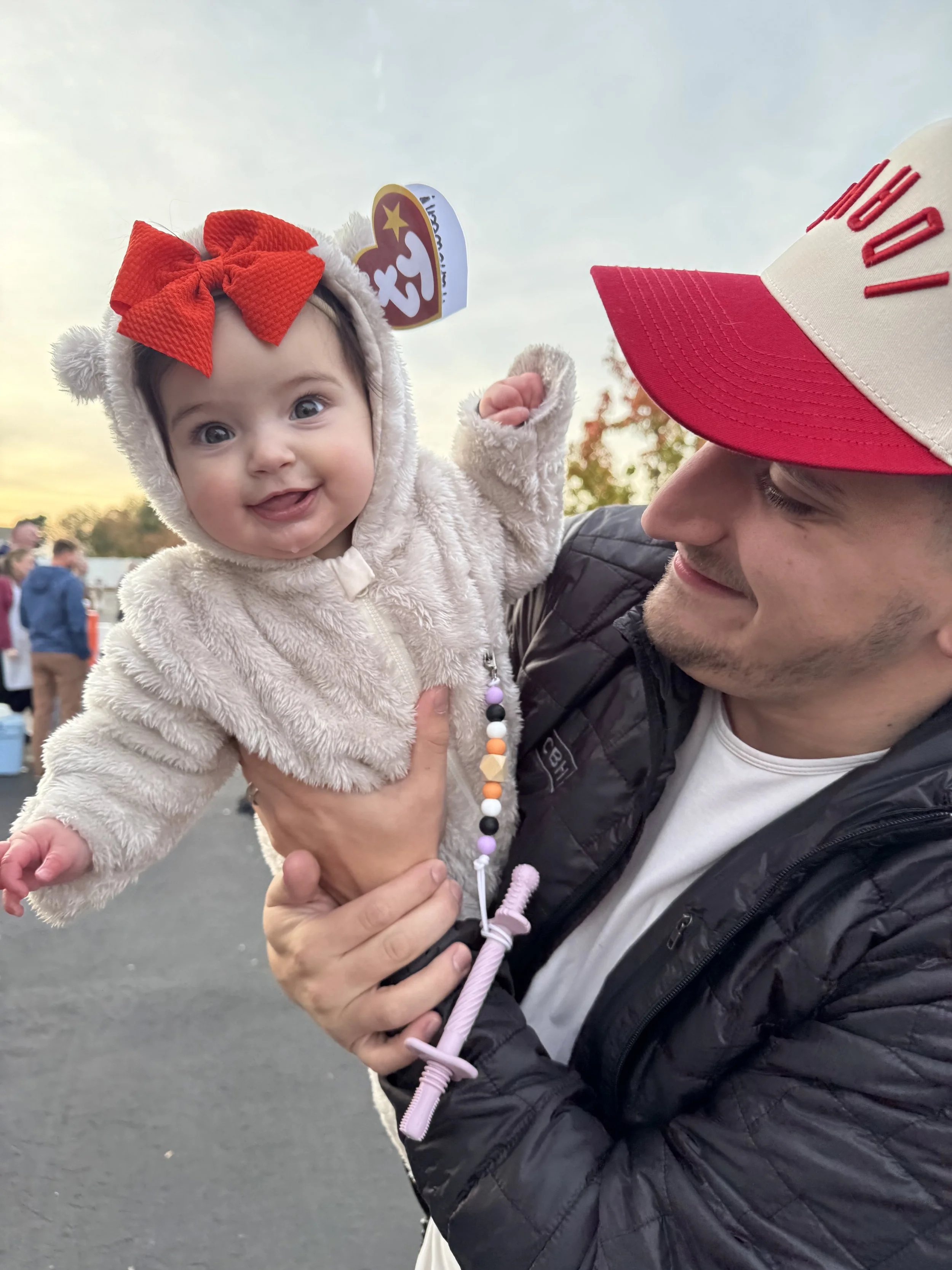 A man holding a smiling baby girl in a bear costume with a red bow and a Beanie Babies tag, outdoors during sunset.