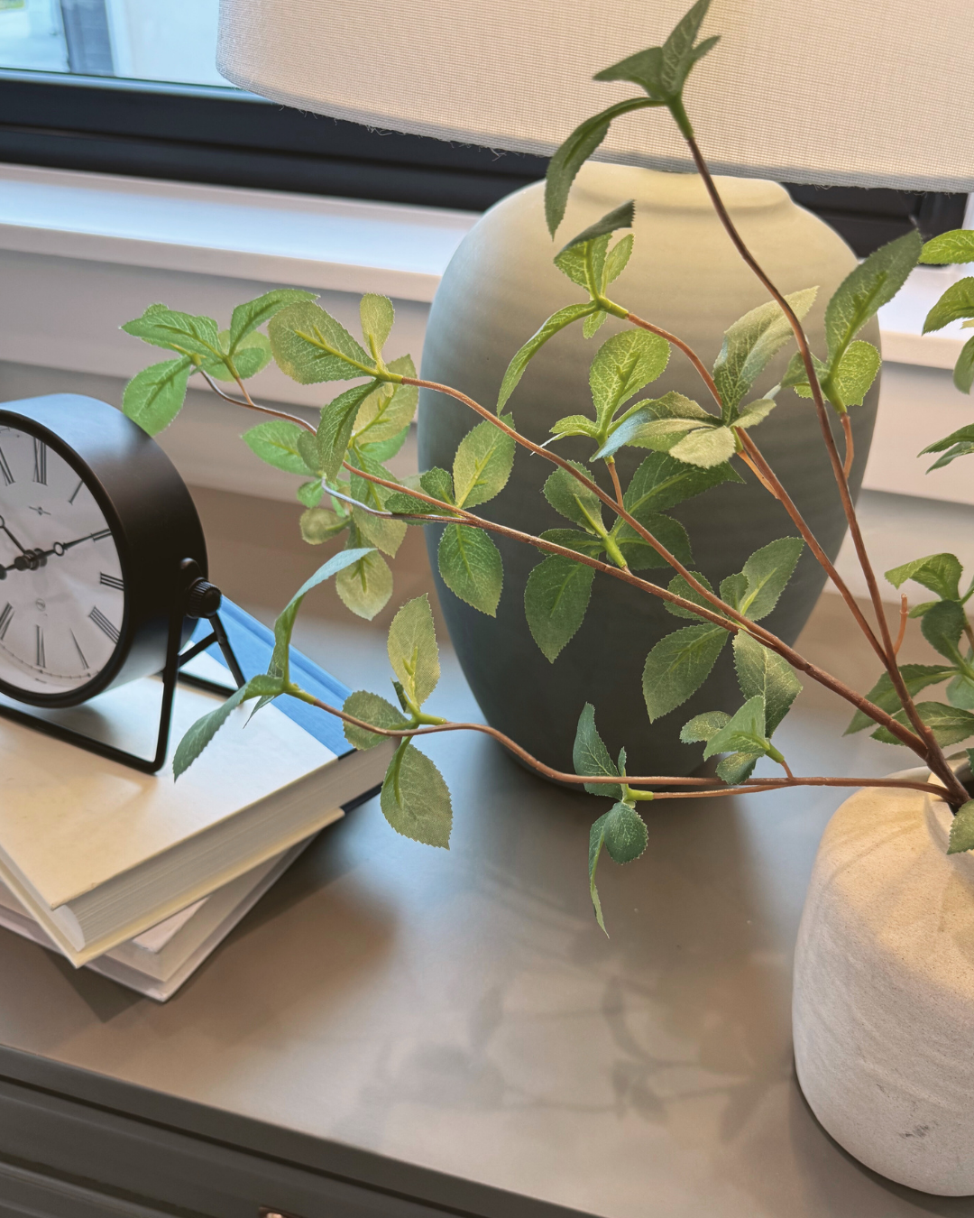 A close-up of a black and white clock, a large green vase with a leafy green plant, a smaller white pot with another plant, and a stack of notebooks near a window.