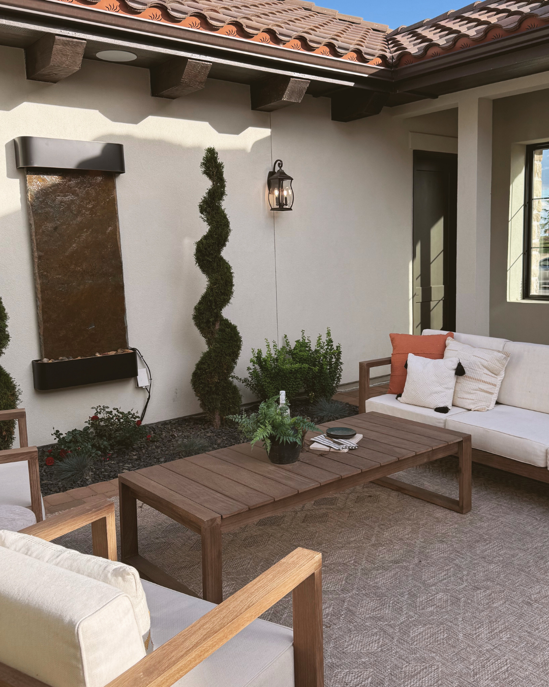 Outdoor patio with white walls, a water feature on the wall, a twisted topiary tree, potted plants, a wall lantern, and seating area with beige cushions and wooden furniture.