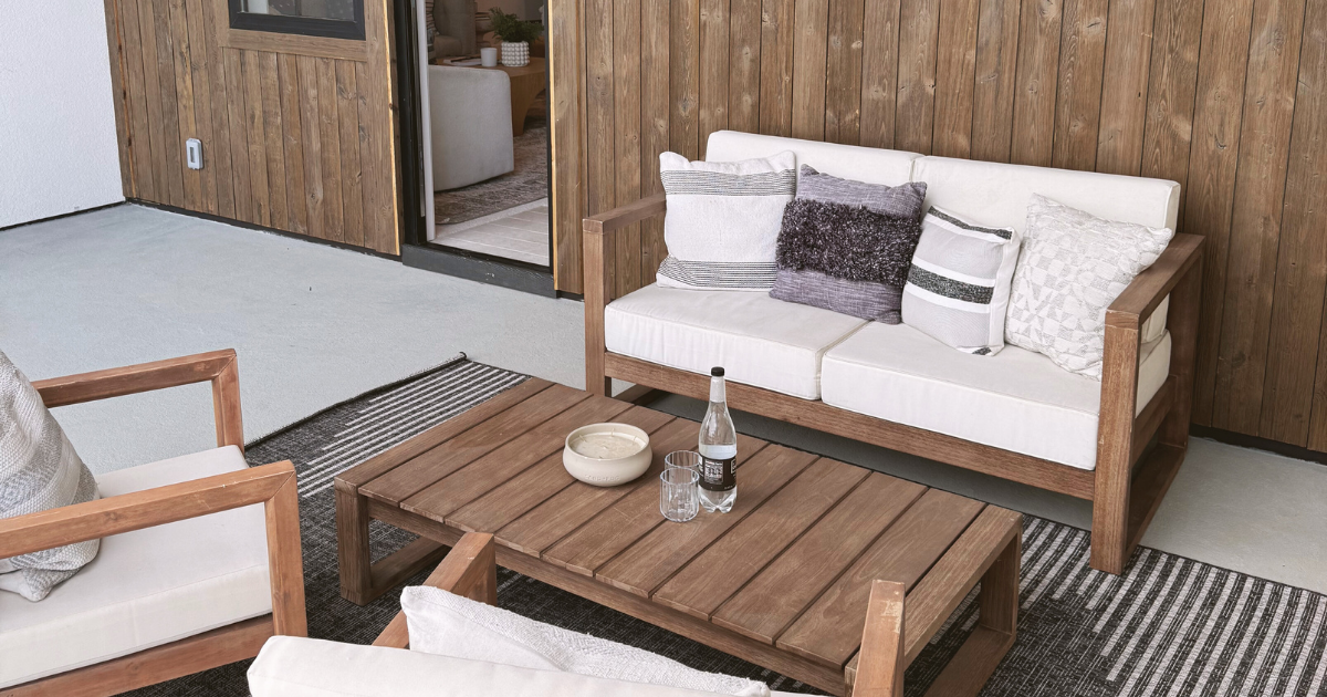 Outdoor patio with wooden furniture, including a sofa with cushions and a coffee table with a bottle, glass, and bowl, next to a wooden wall and an interior room visible through a sliding door.