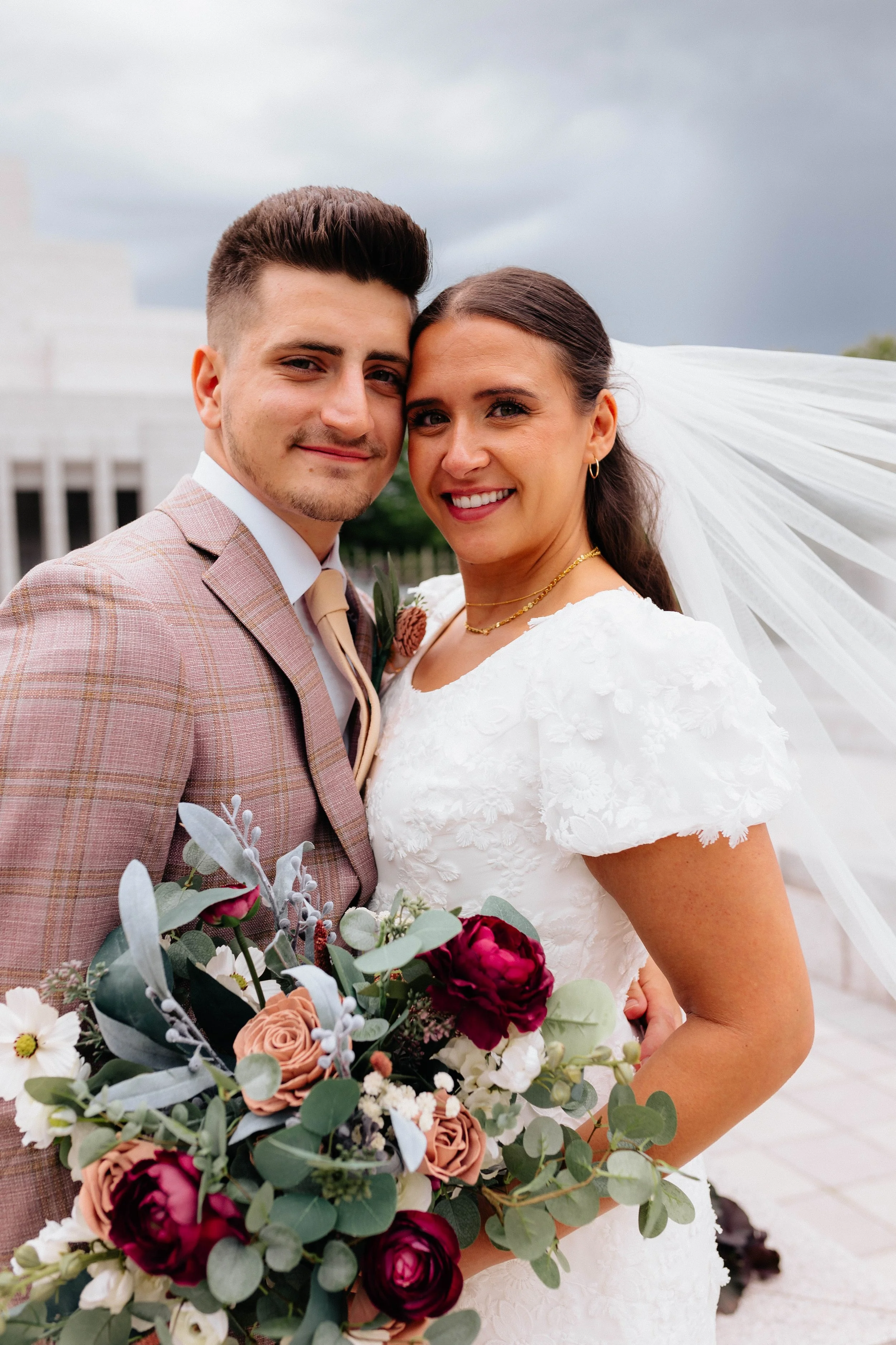 A bride and groom smiling and embracing outdoors, the bride holding a bouquet of flowers, with a cloudy sky in the background.