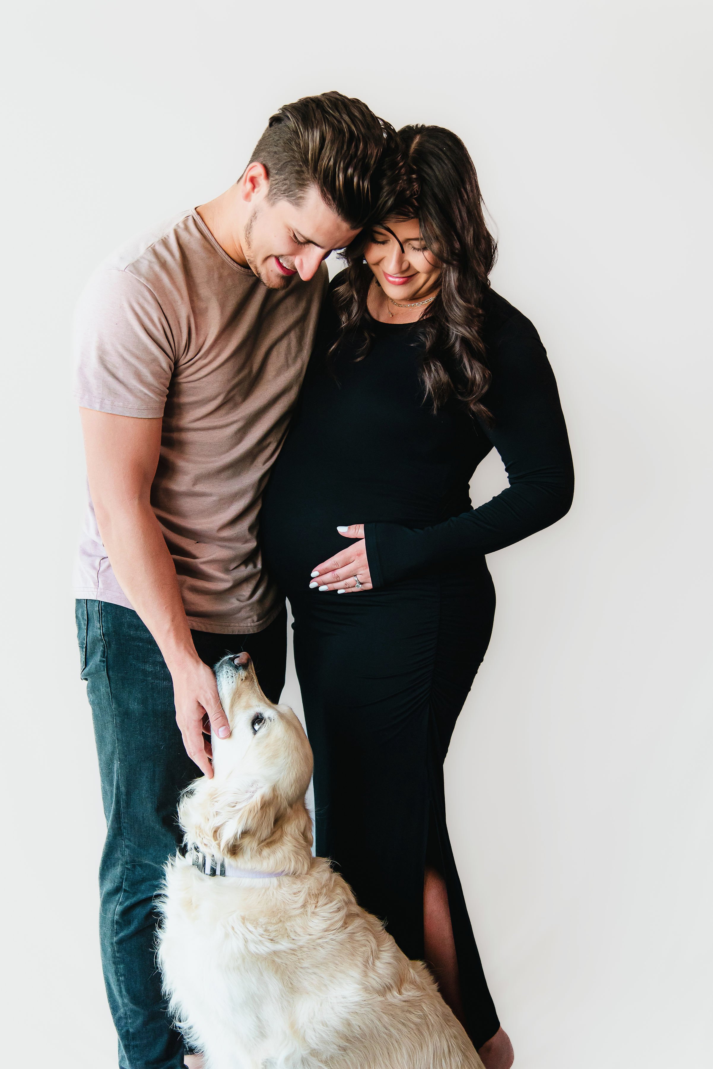 A pregnant woman and a man with a golden retriever puppy against a plain white wall.