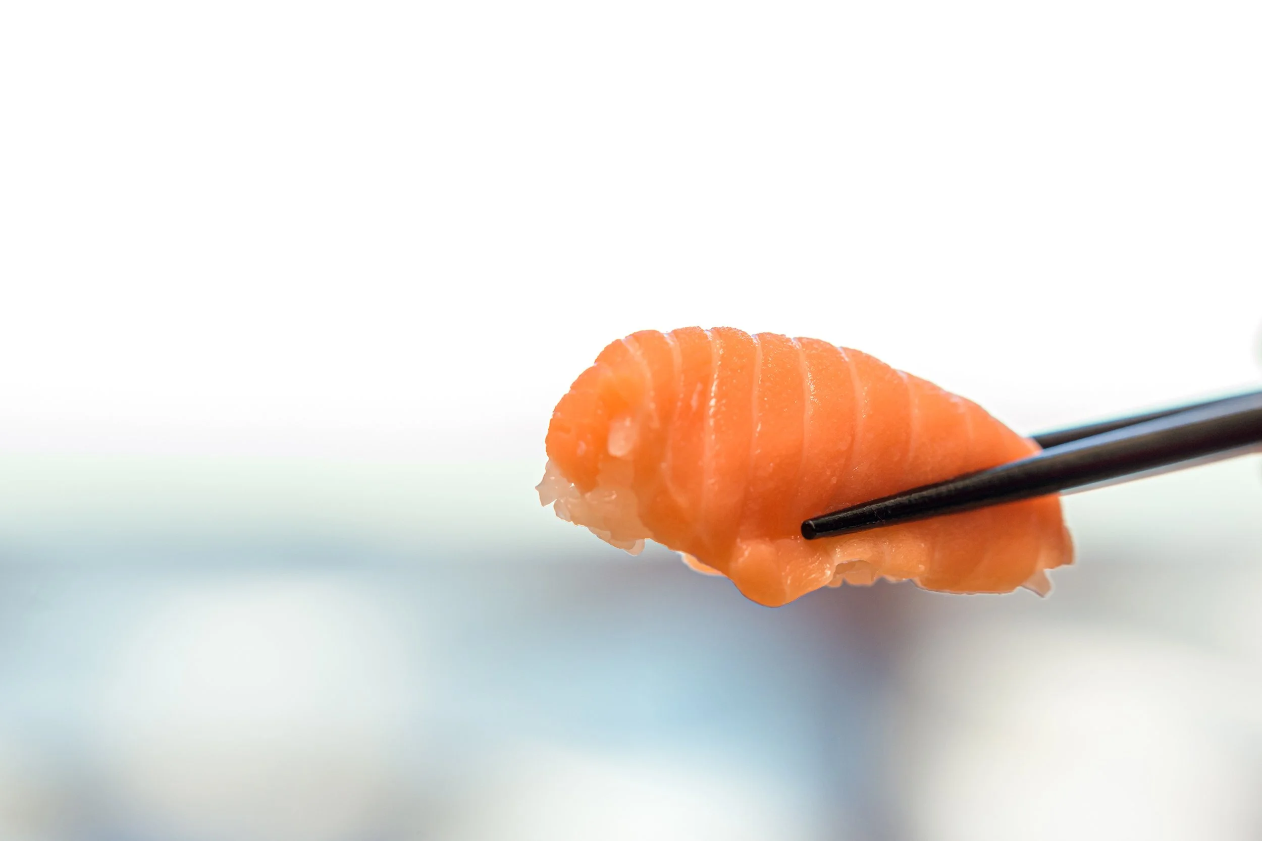A piece of fresh salmon sashimi held with black chopsticks against a light, blurred background.