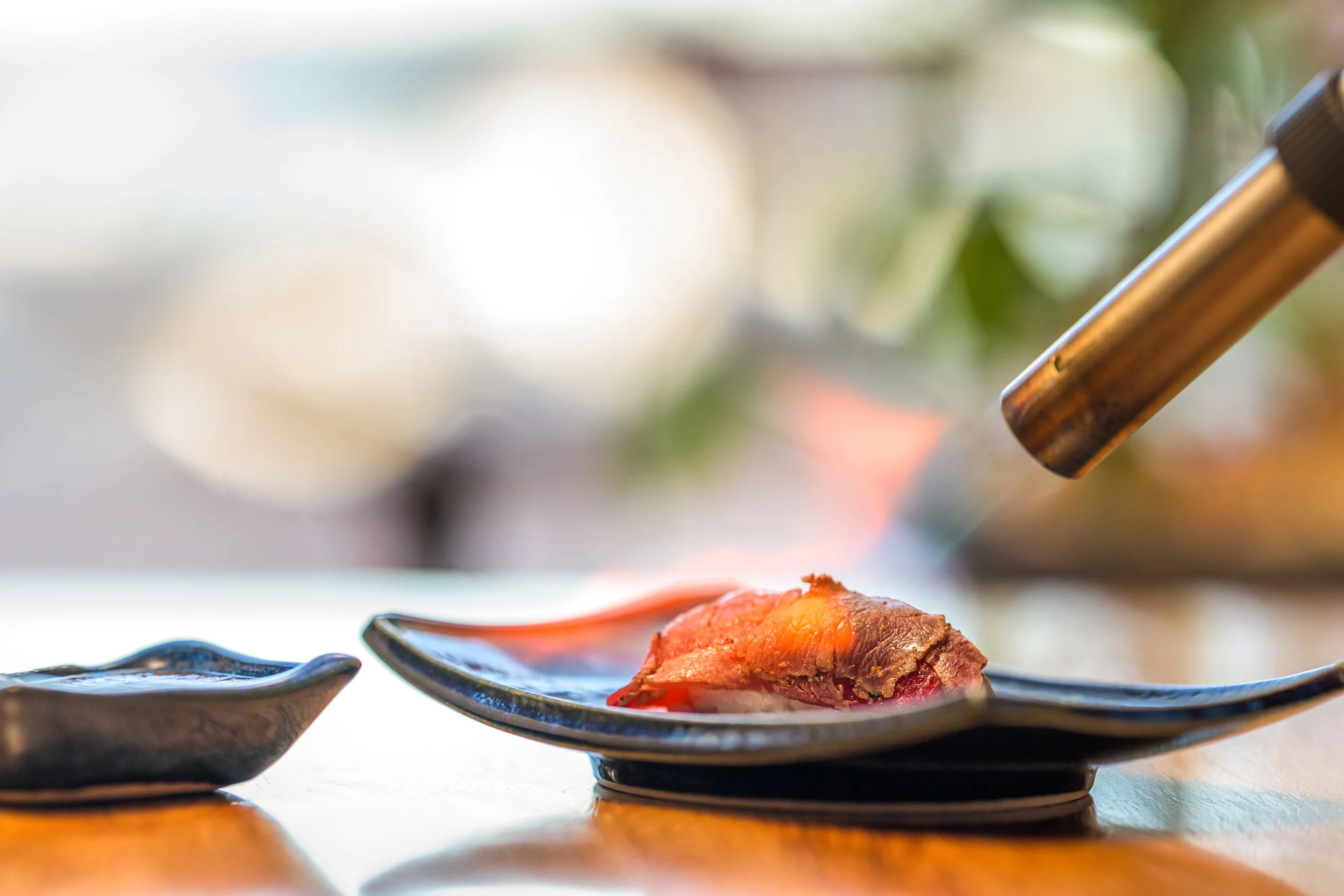 Close-up of a piece of sushi on a decorative black plate, with a blowtorch pointed at it, in a restaurant setting.