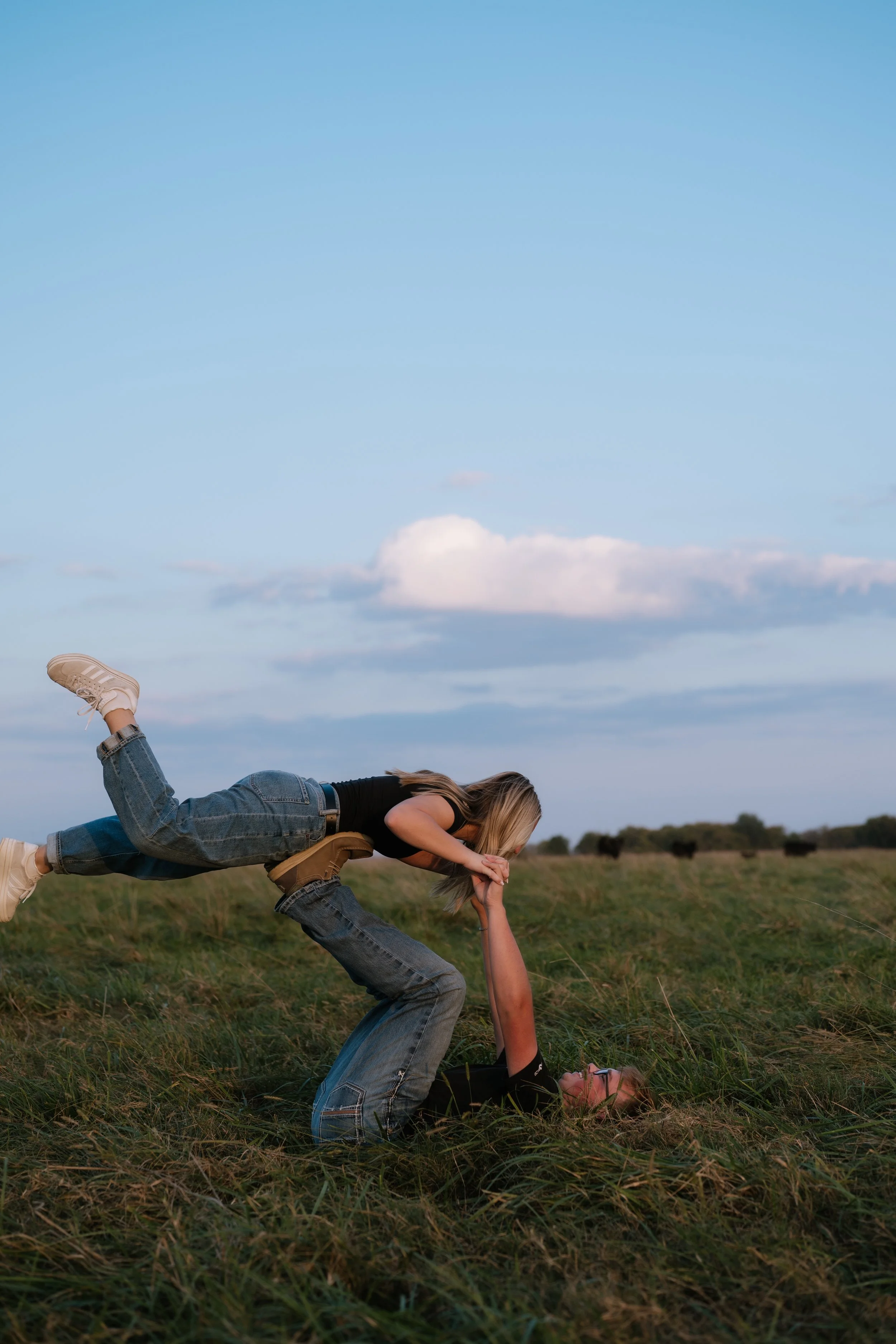 Two women playing on a grassy field during daytime, with one lying on the ground and the other on top, holding hands and engaged in playful activity. couple photographer portrait photographer Kansas photographer wedding photographer