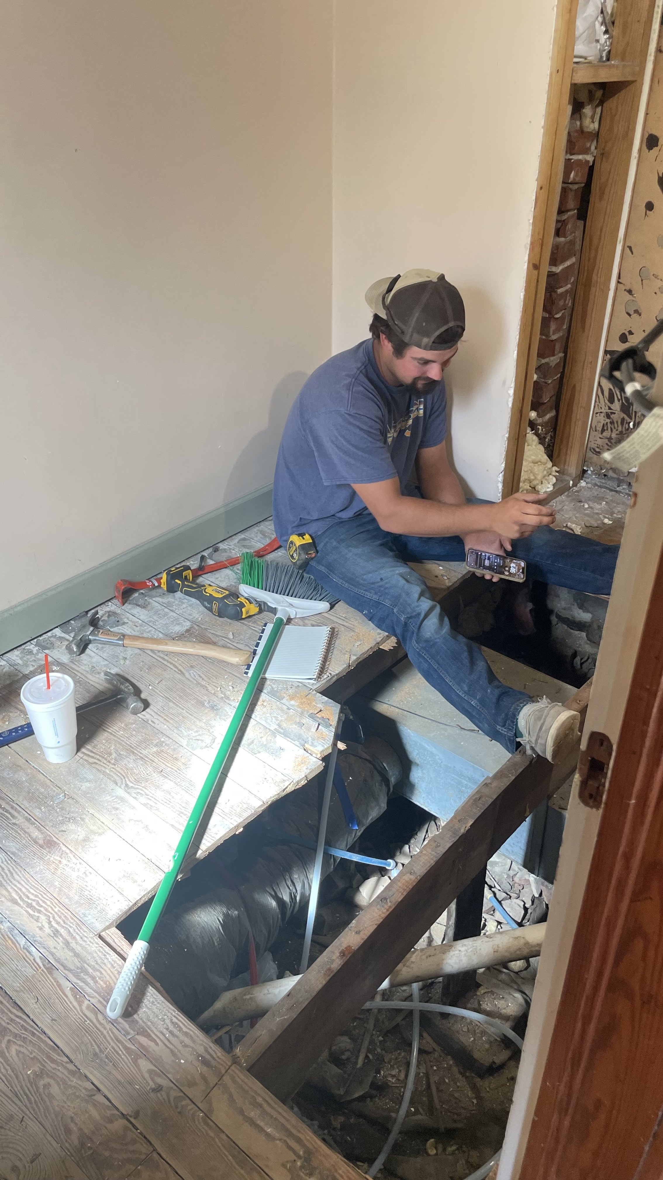 A man sitting on the edge of a house floor under renovation, using a phone, surrounded by tools and construction materials.