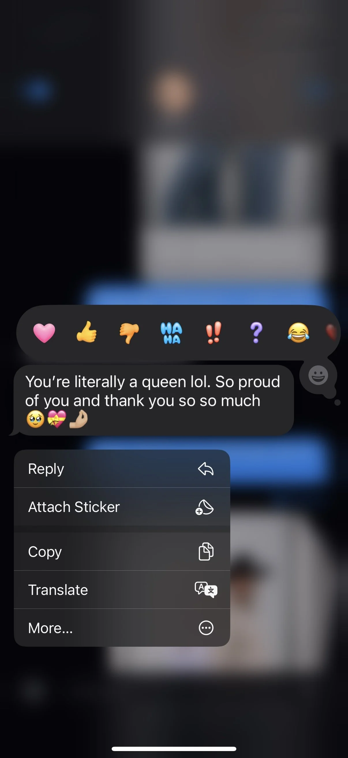 A blurry screenshot of a social media message conversation with a background of a person sitting at a desk, the message reads: "You're literally a queen lol. So proud of you and thank you so so much" with emojis, and a menu of options including reply, attach sticker, copy, translate, and more.