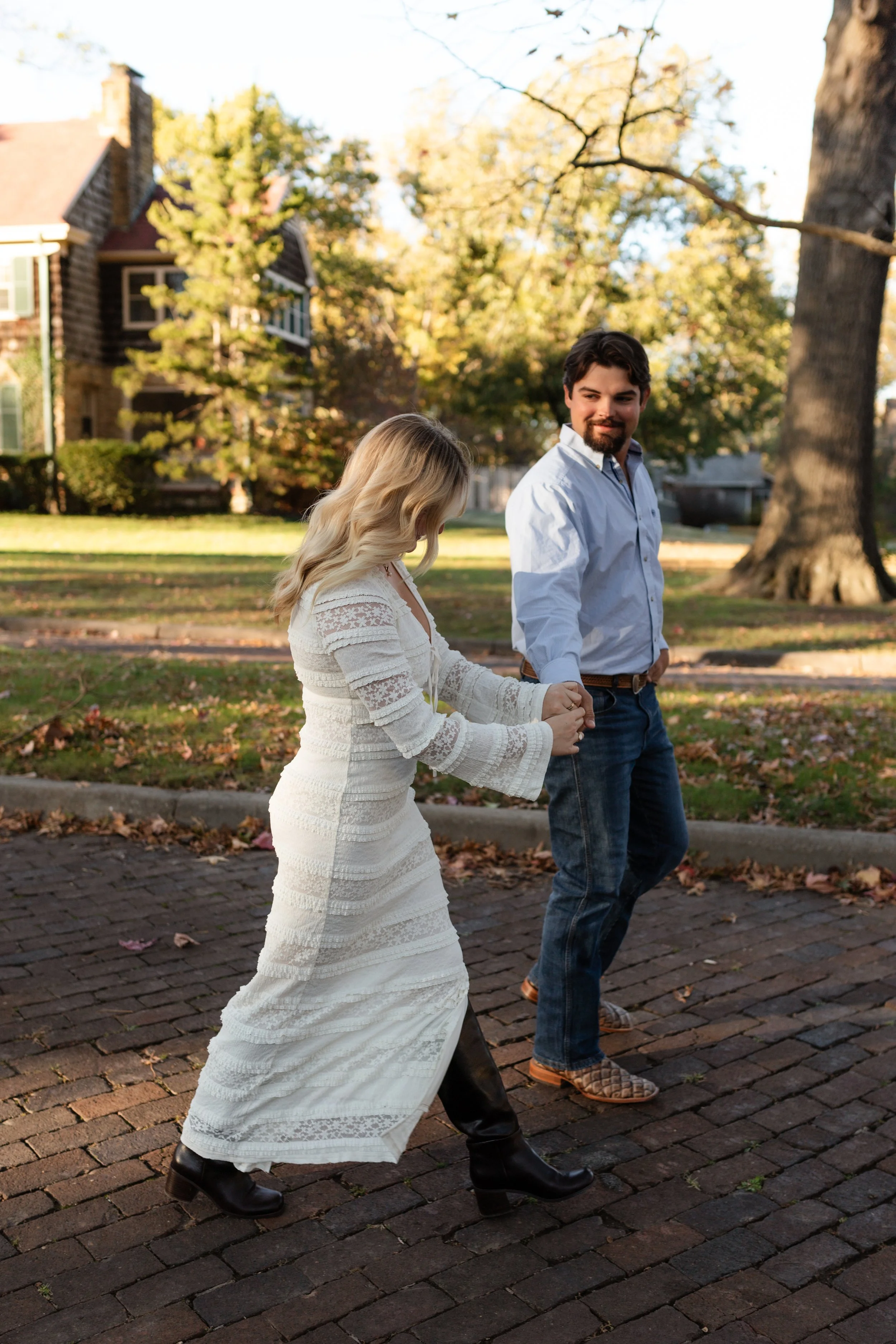 A couple holding hands and walking on a brick sidewalk in a park with fallen leaves and large trees during fall. Joplin Missouri engagement photoshoot