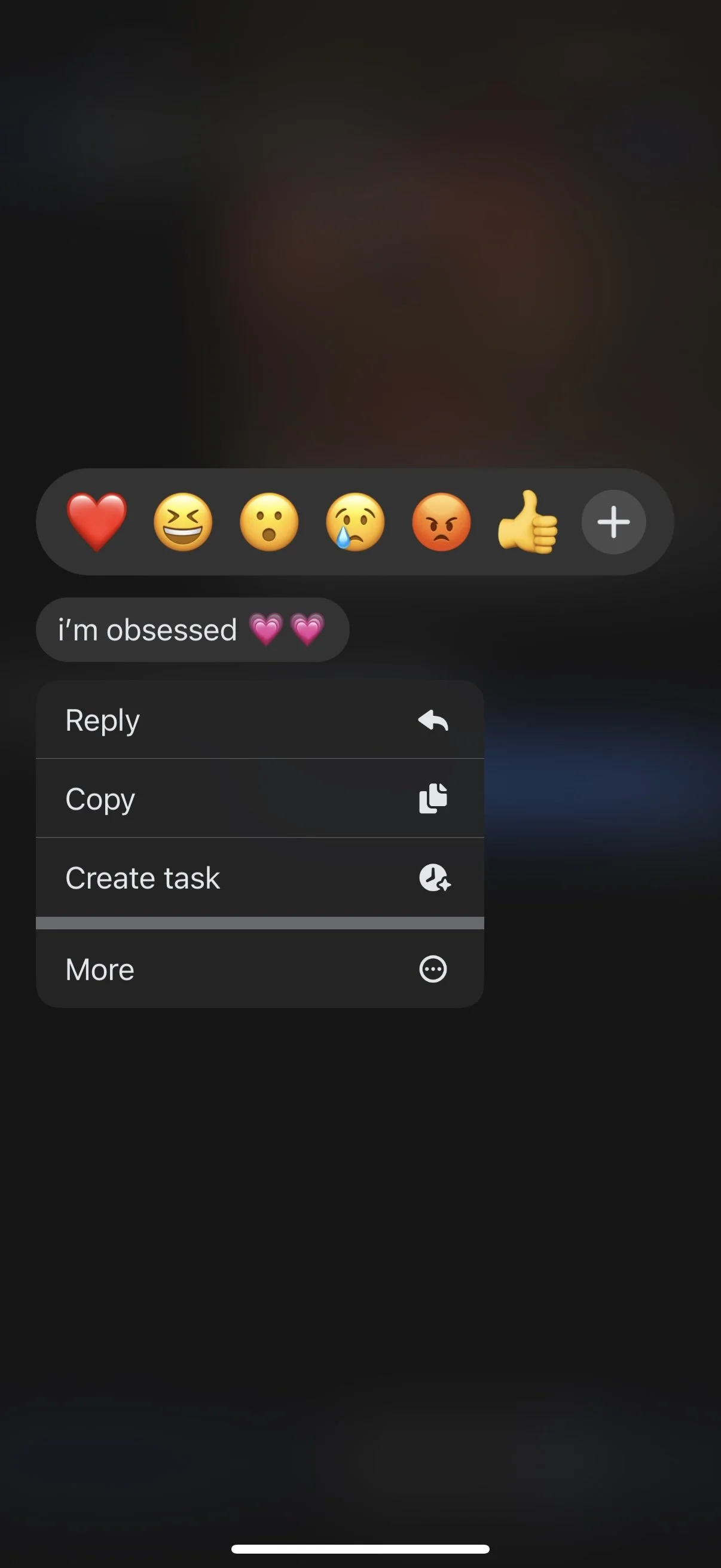 A screenshot of a social media post with a dark background, showing a series of emojis including a red heart, laughing face, surprised face, crying face, angry face, and thumbs up. Below the emojis, there is a caption that says, "i'm obsessed" with two pink hearts. The screenshot also displays a menu with options: reply, copy, create task, and more.