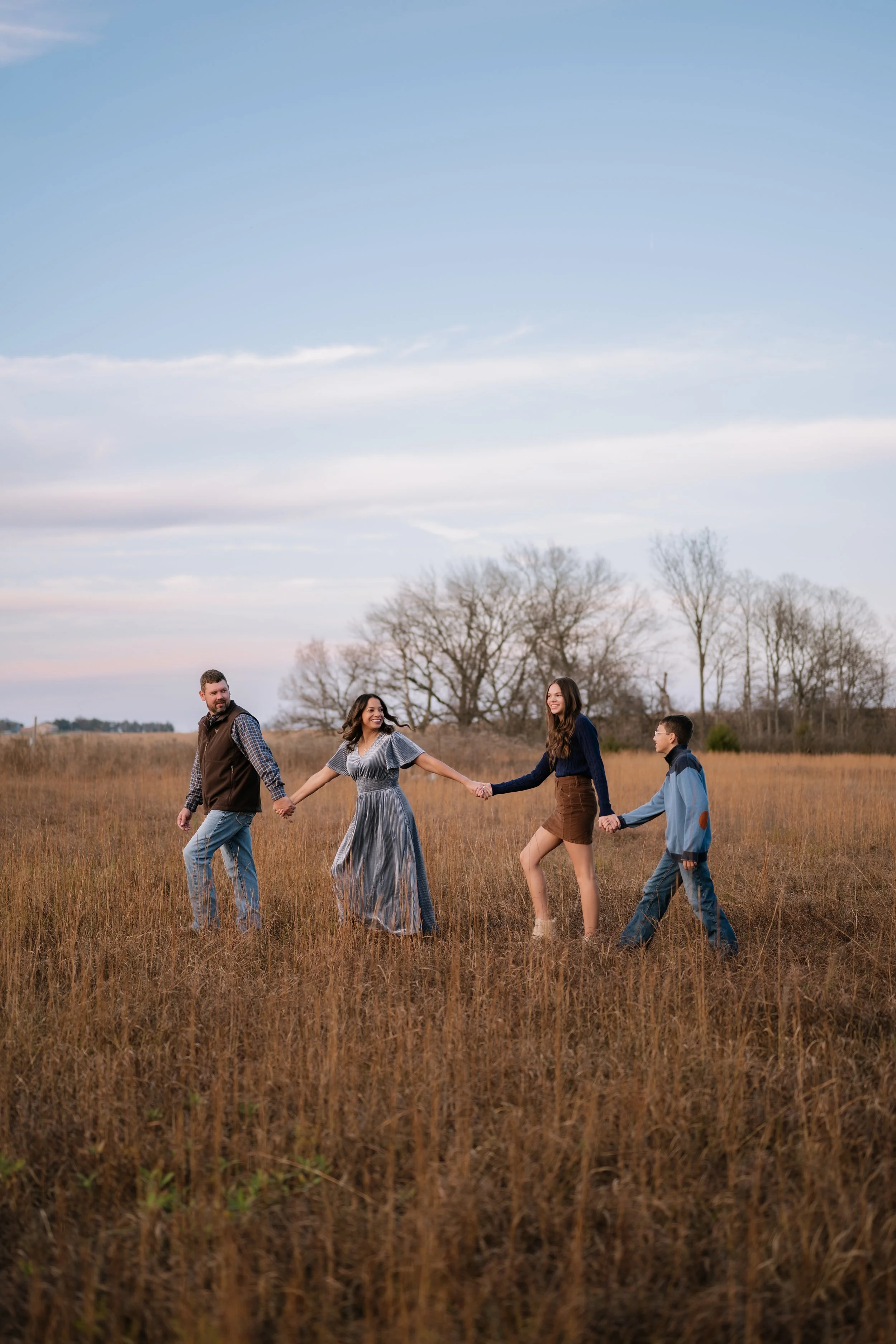 A family of four holding hands and walking through a grassy field during sunset, with trees in the background. Fredonia Kansas family photography.