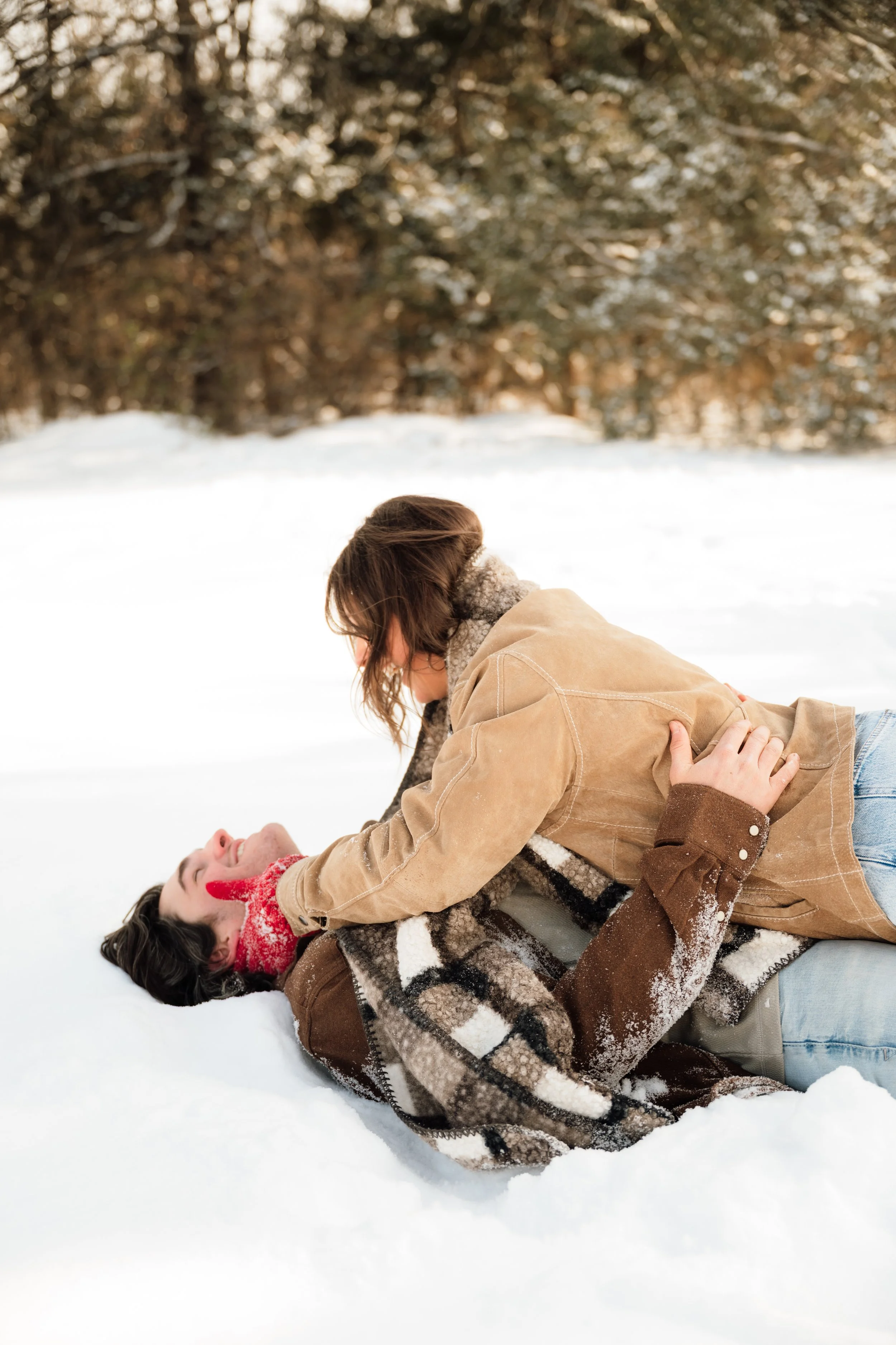 Couple playing in the snow, one lying on the ground and the other leaning over them, with snow-covered trees in the background. Kansas photographer