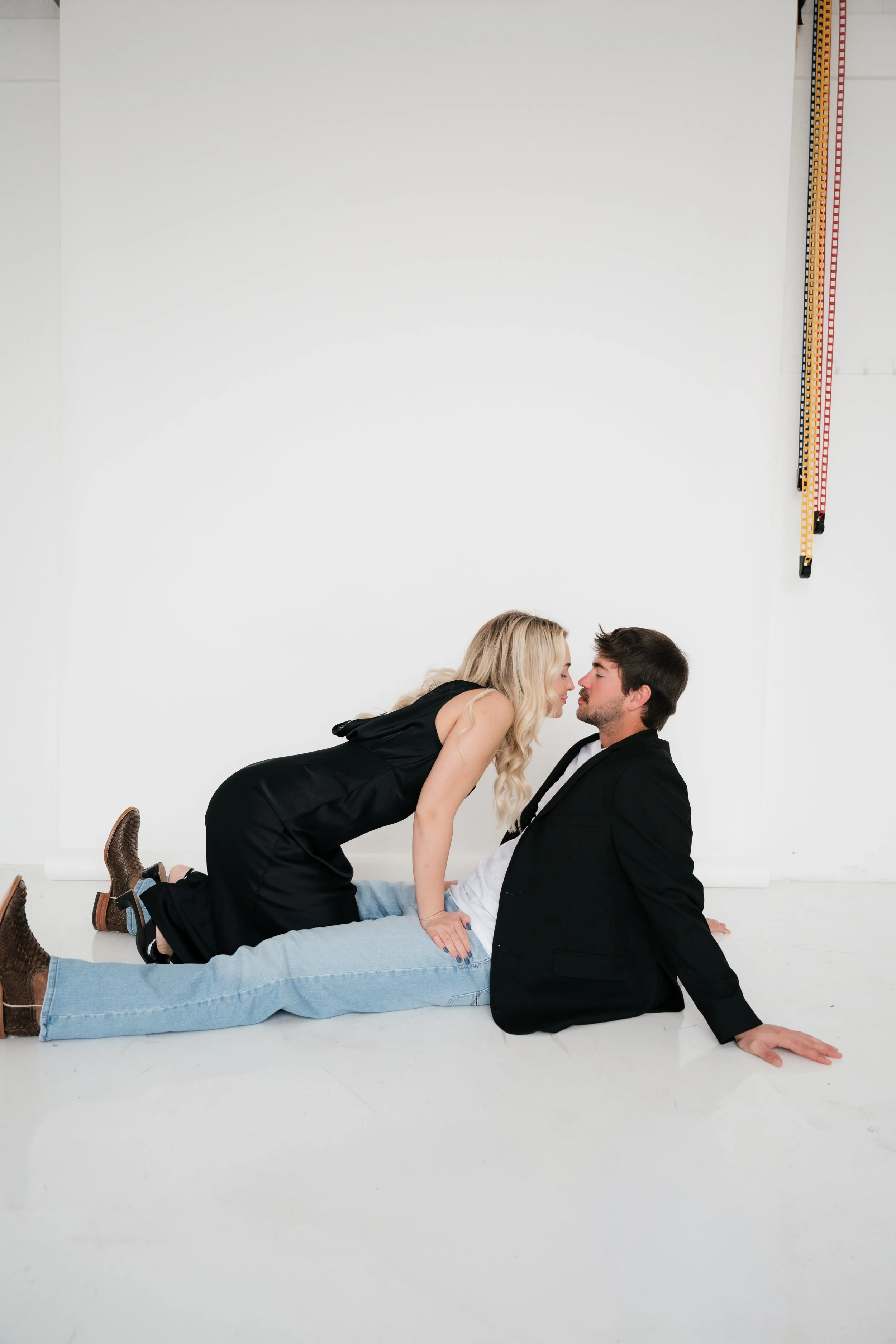 A couple on the ground, leaning close and about to kiss, in a minimalist studio with a white background.
