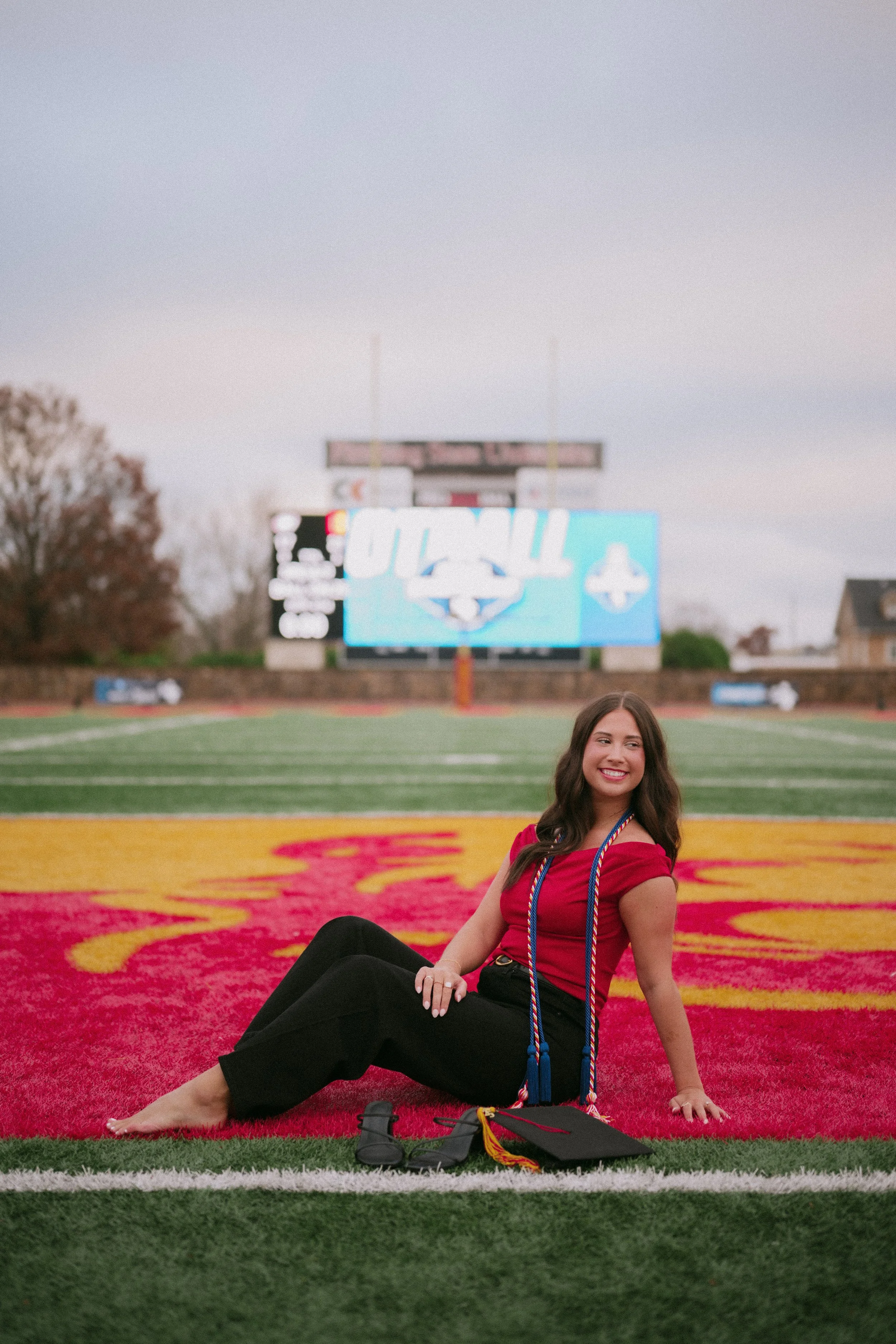 A young woman in a red top and black pants sitting on a football field with graduation cords, a diploma, and cap nearby, smiling with a scoreboard and football field in the background.