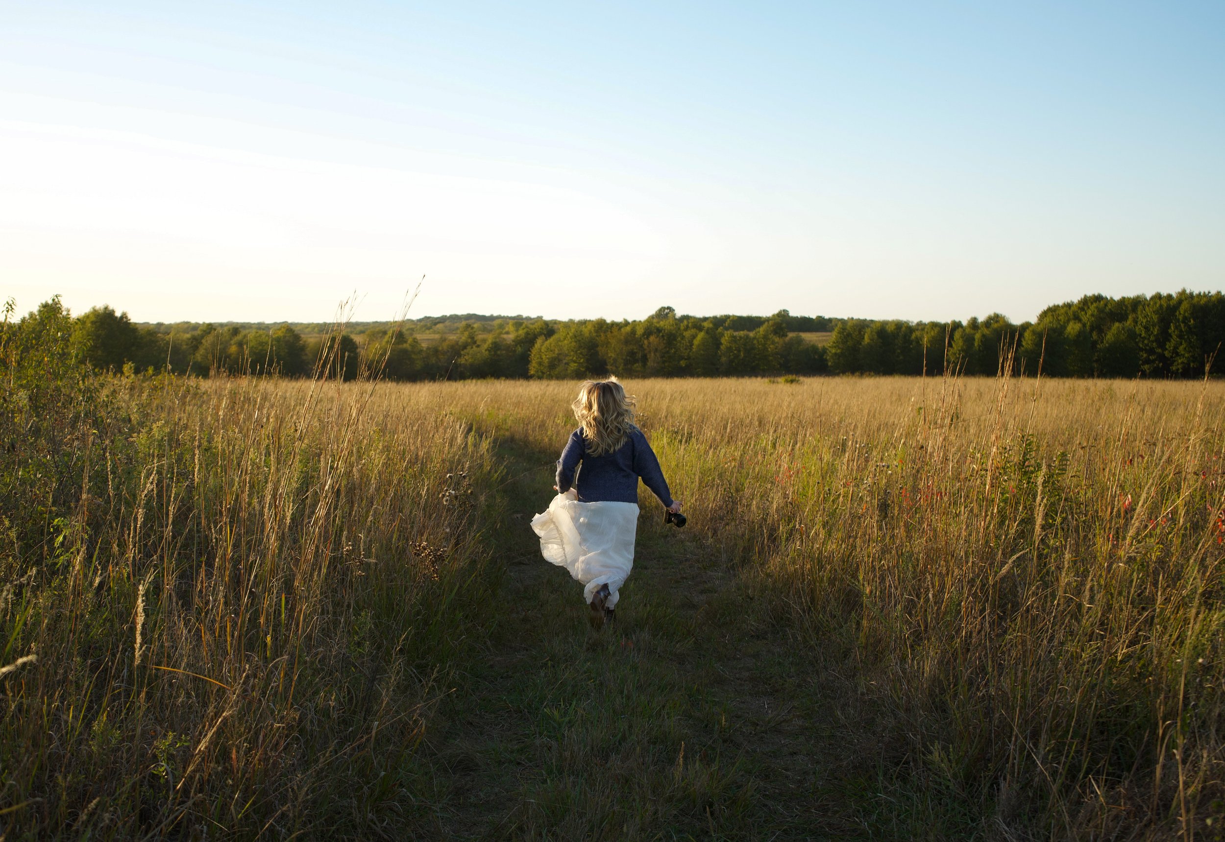 A girl with blonde hair walking on a dirt path through a grassy field in a rural area during late afternoon or early evening. Midwest photographer Kansas photographer Missouri photographer Pittsburg wedding