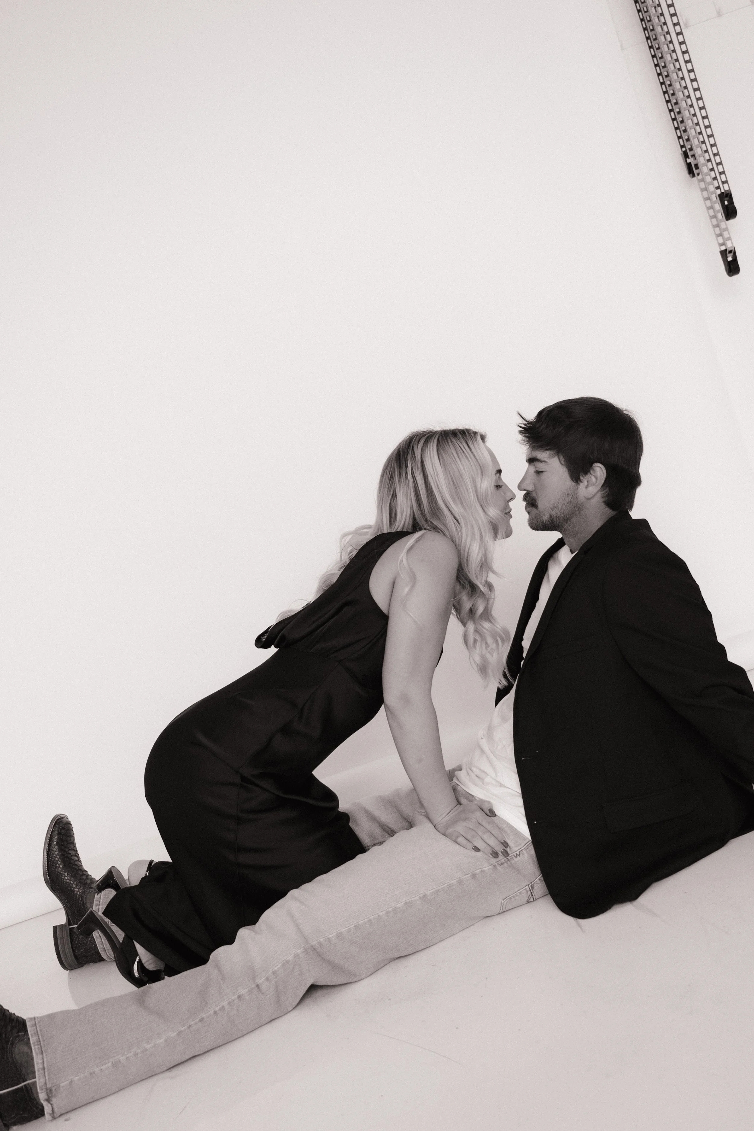 A couple on the ground, leaning close and about to kiss, in a minimalist studio with a white background.