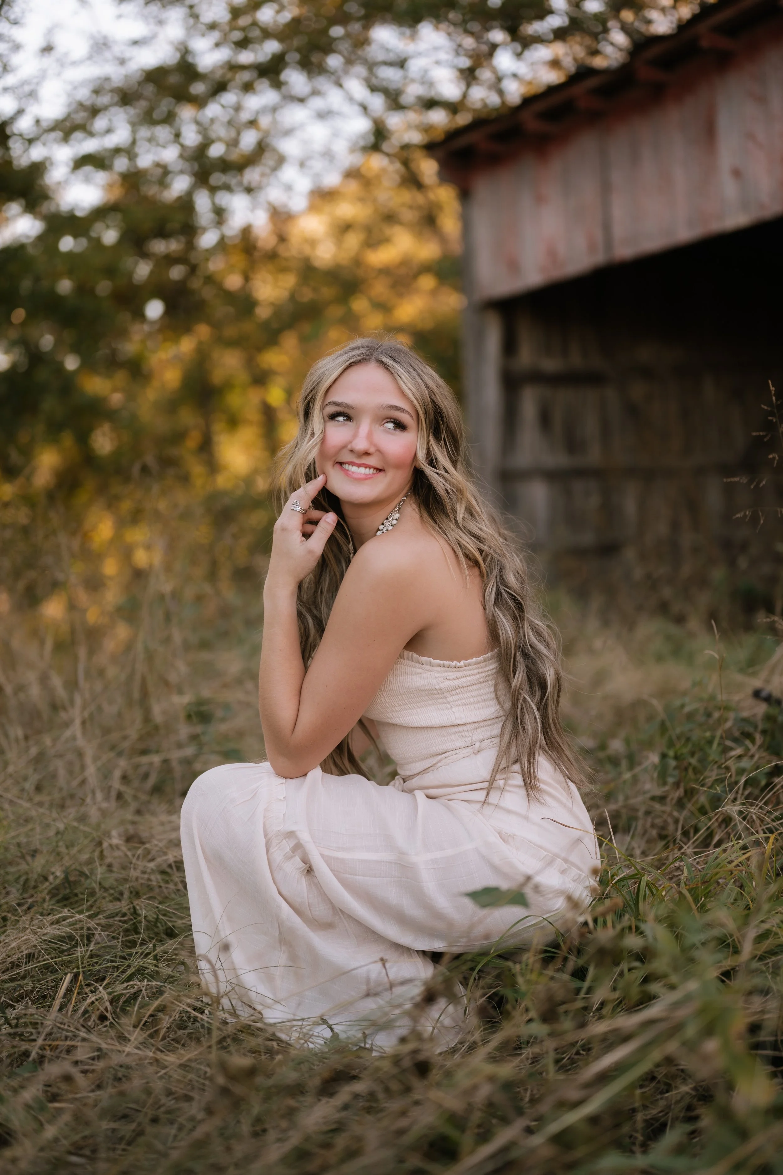 A young woman with long wavy blonde hair smiling and looking away, sitting in tall grass near an old wooden building in a natural outdoor setting during fall.