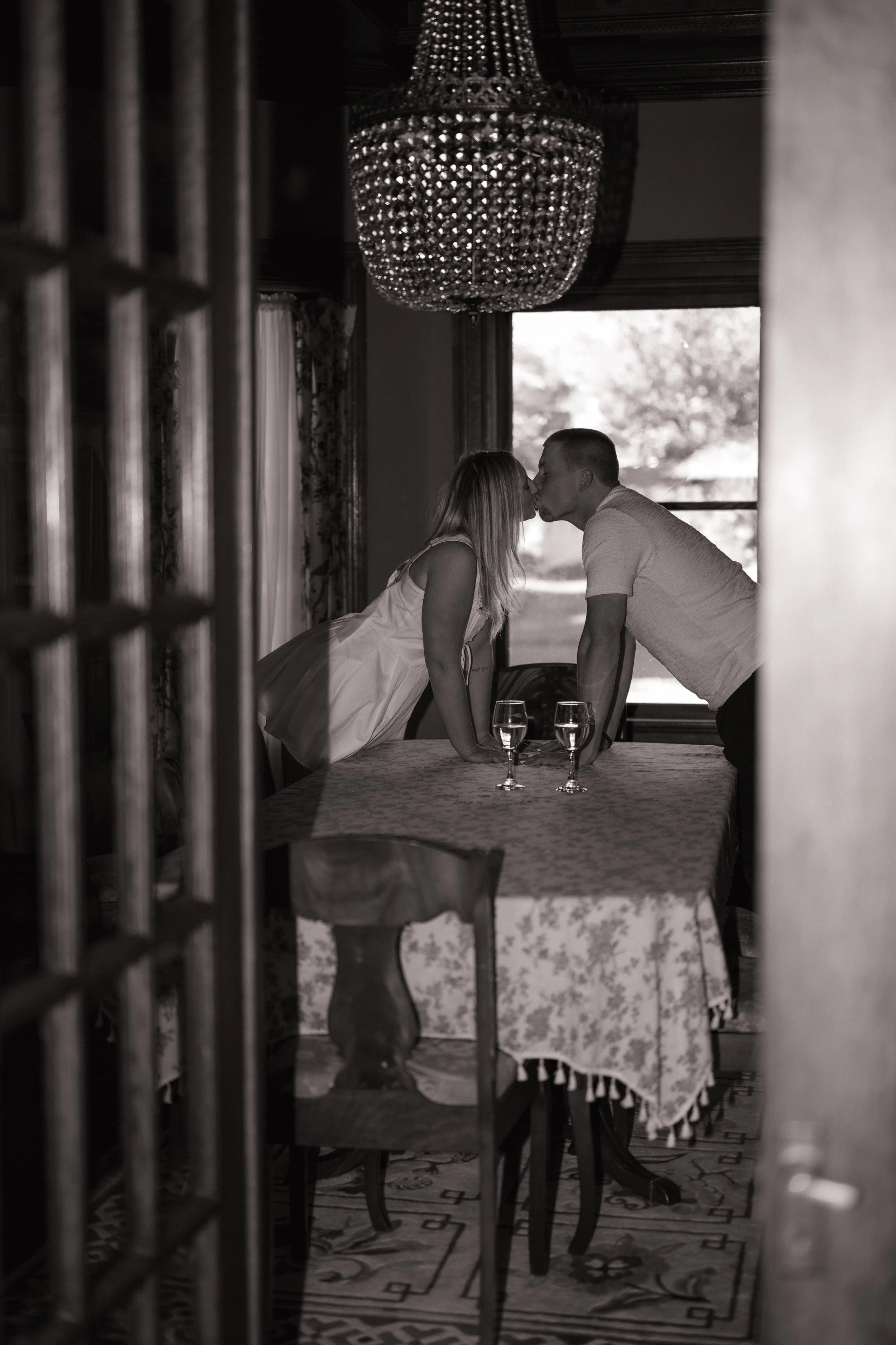 A couple leaning over a dining table, about to kiss, with two glasses of water on the table, seen through an ornate wooden door frame.