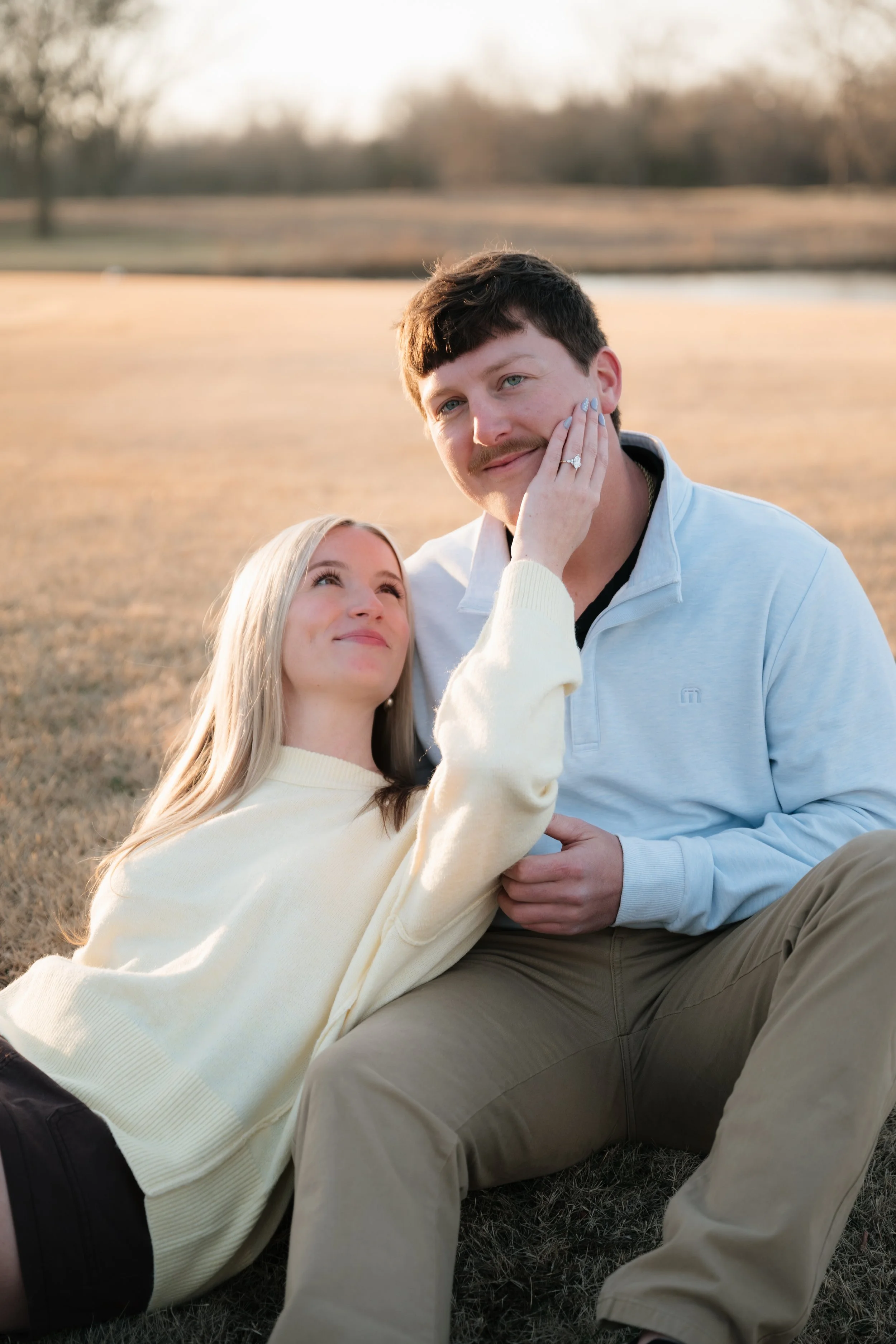 A woman gently touches a man's face as they sit outside on a grassy field during sunset, with trees in the background. Kansas City engagement photographer
