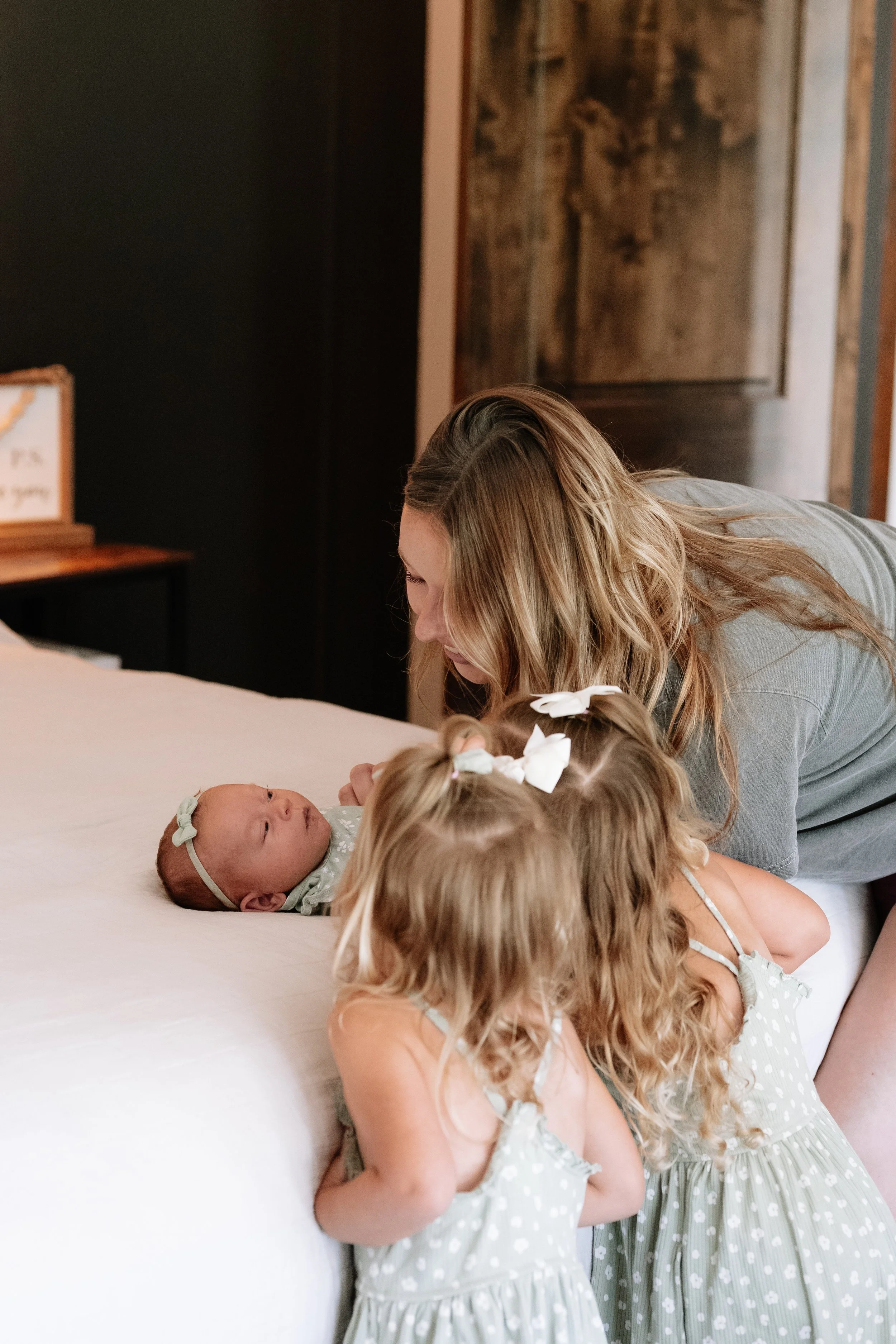 A woman leans over a bed, interacting with a baby girl lying on the bed, while two young girls with curly hair and matching polka dot dresses observe.