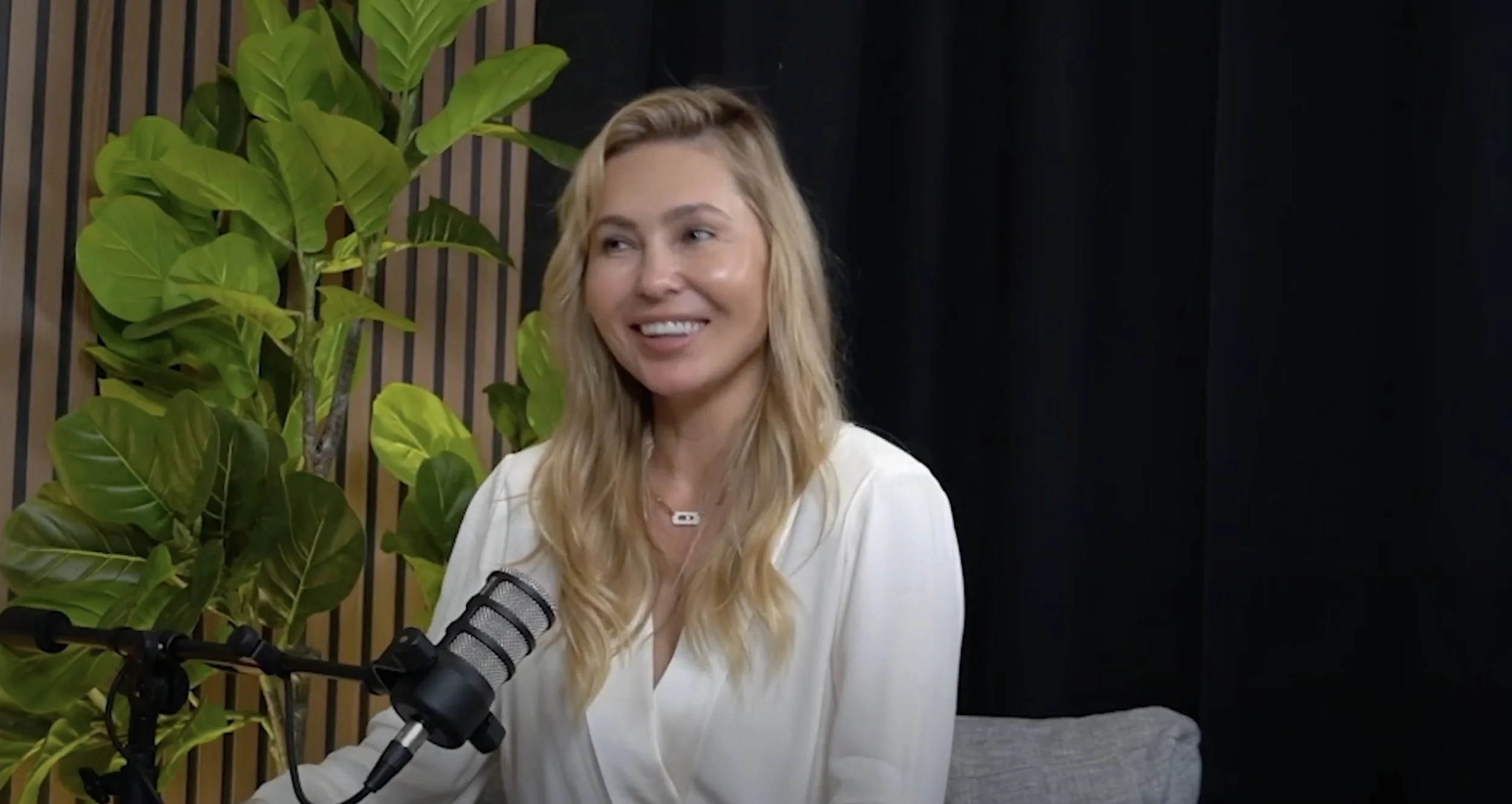 A woman with blonde hair smiling while sitting in front of a microphone, with green plants and a wooden slat wall in the background.