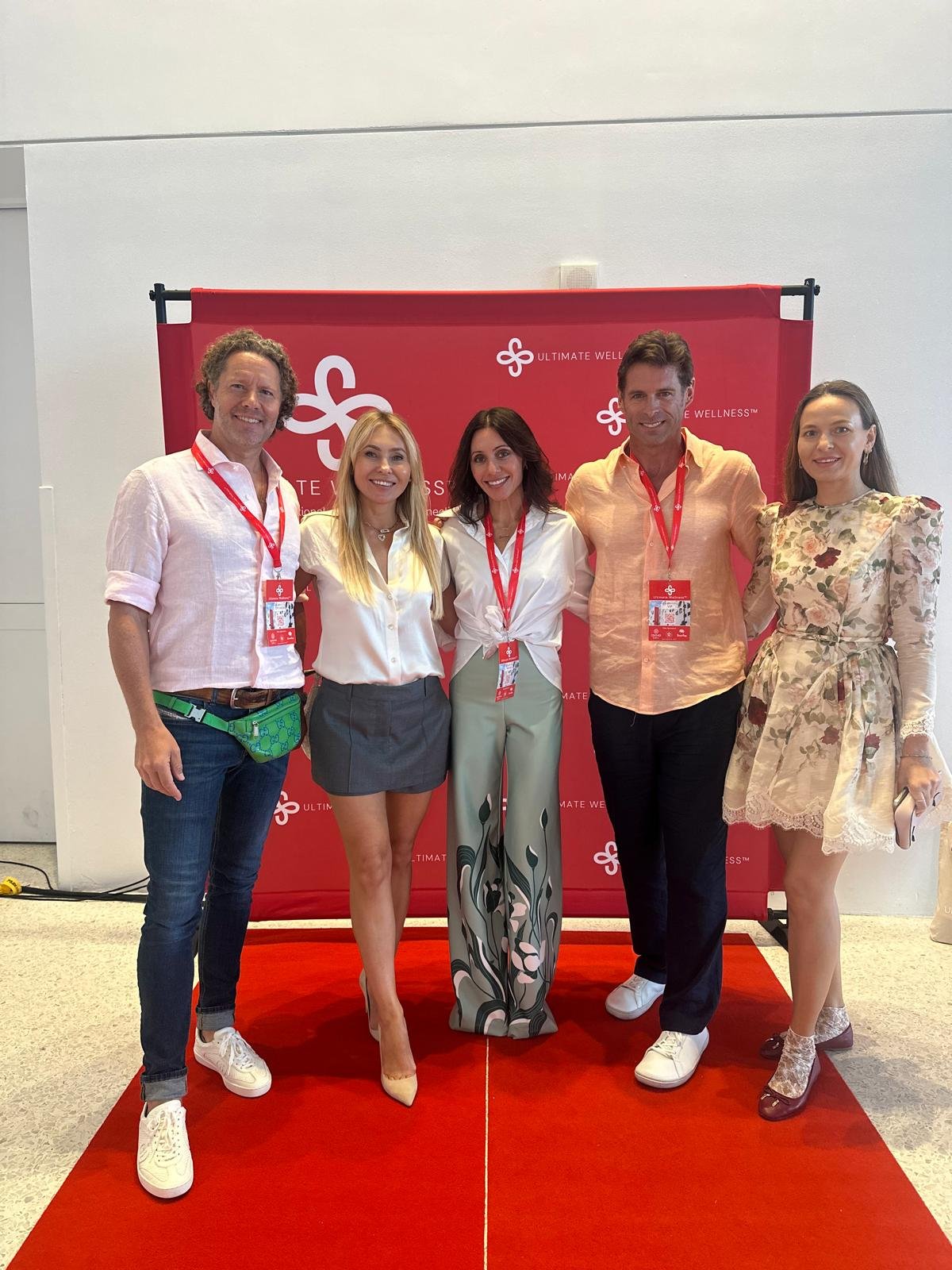 A group of five people standing together on a red carpet, smiling at the camera, with a corporate backdrop behind them. The backdrop is red with white text and logos that say "Ultimate Wellness."
