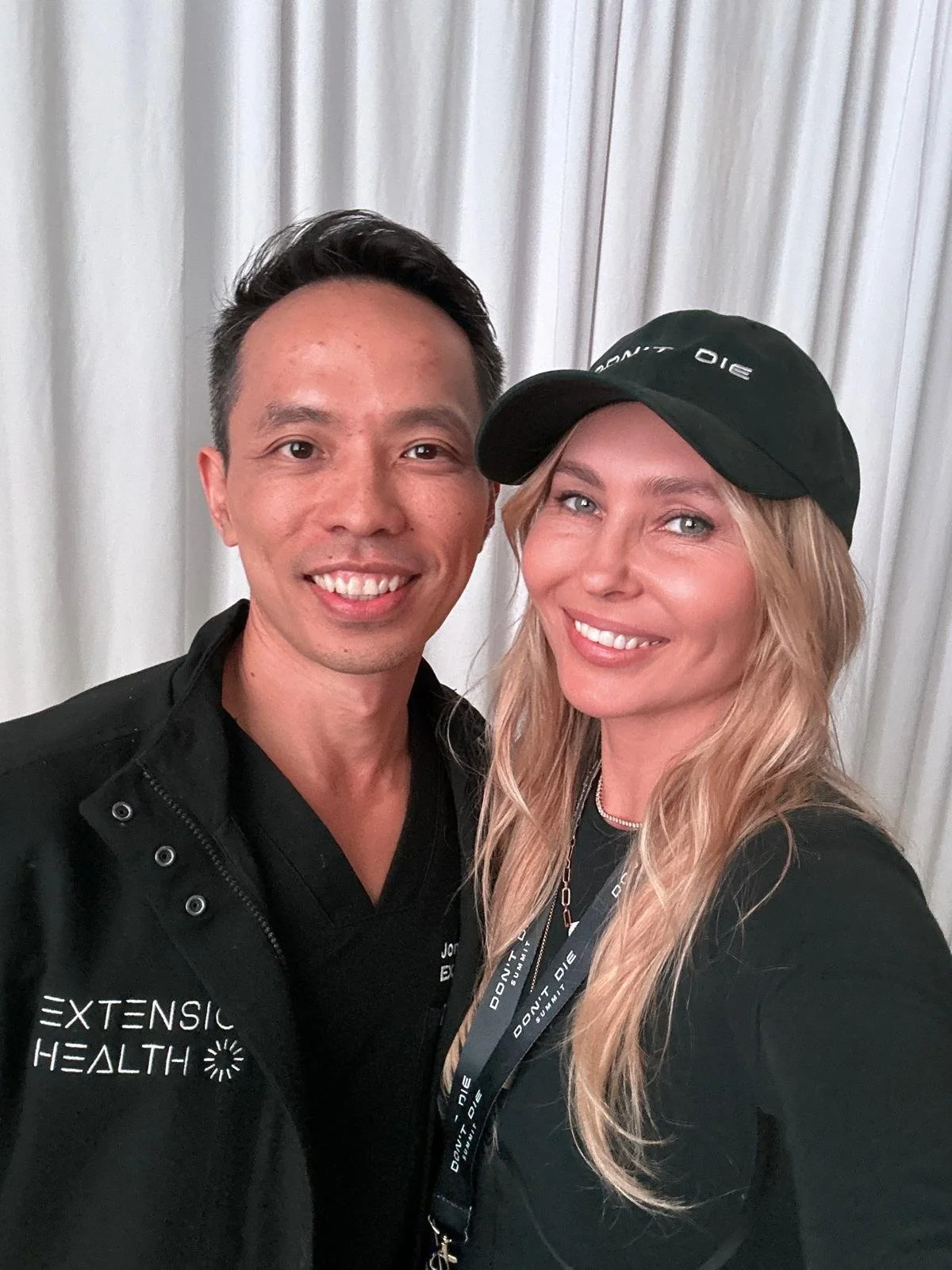 A man and woman posing together for a photo, both smiling. The man is wearing a black jacket with 'EXTENSIVE HEALTH' written on it, and the woman is wearing a black hat and a black shirt, with a lanyard around her neck.